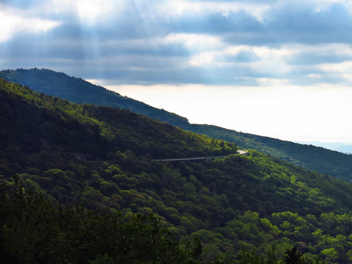 Winding road on a lush green mountain slope with sun rays breaking through clouds.