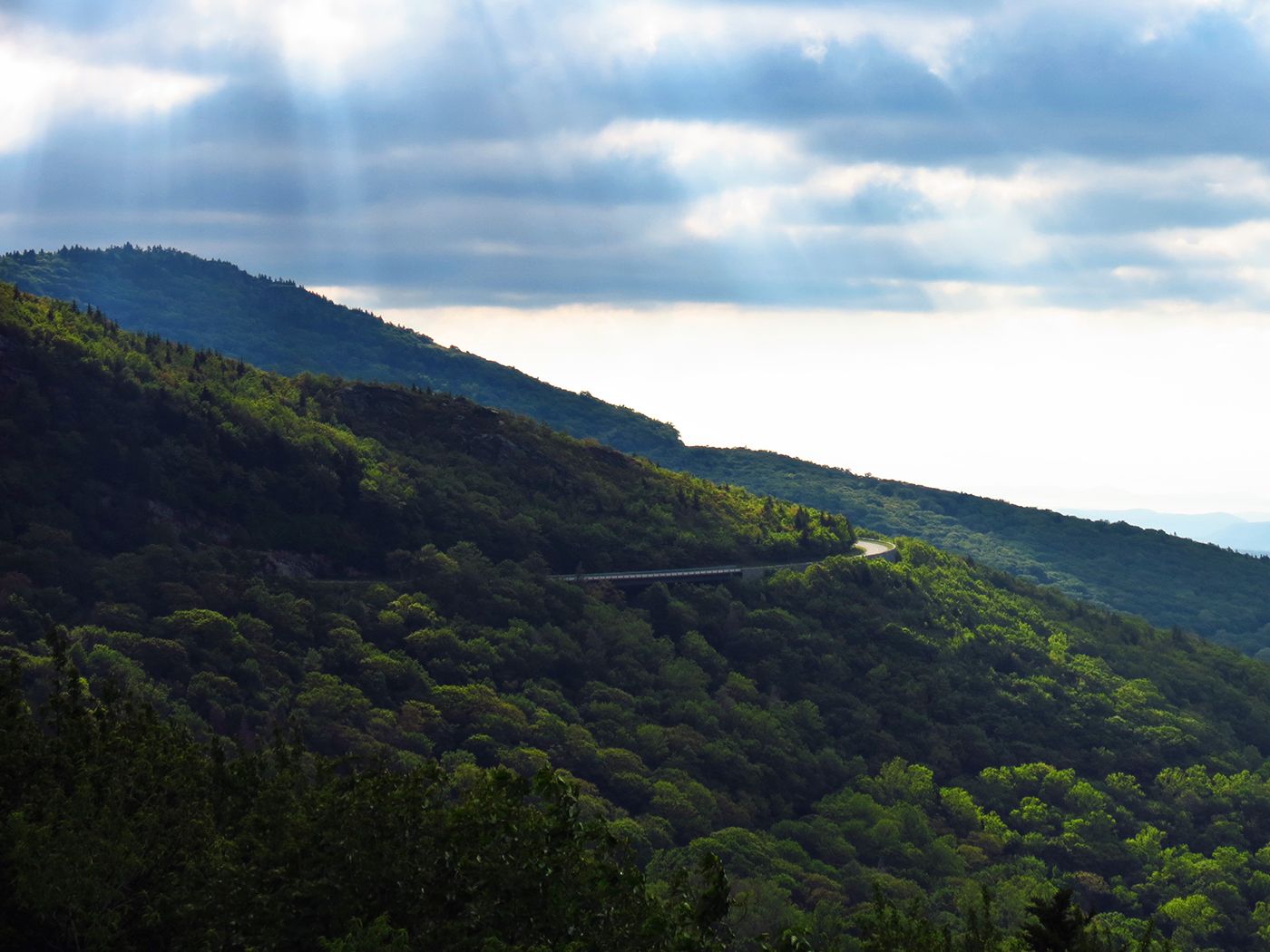 Winding road on a lush green mountain slope with sun rays breaking through clouds. - Rough Ridge Overlook – Pilot Ridge Overlook loop from Rich Mountain