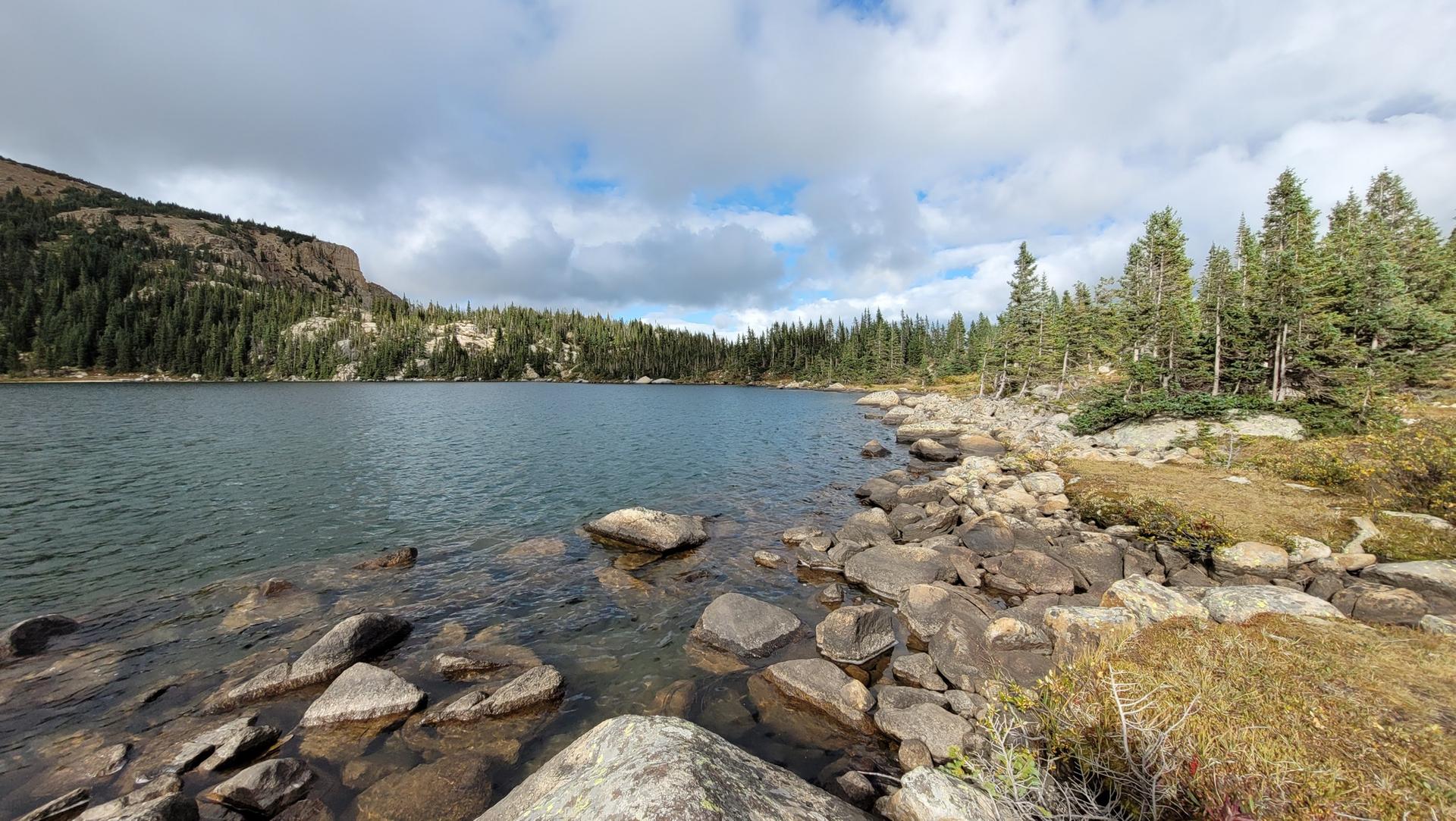 A serene mountain lake with a rocky shore and pine forest under a cloudy sky.