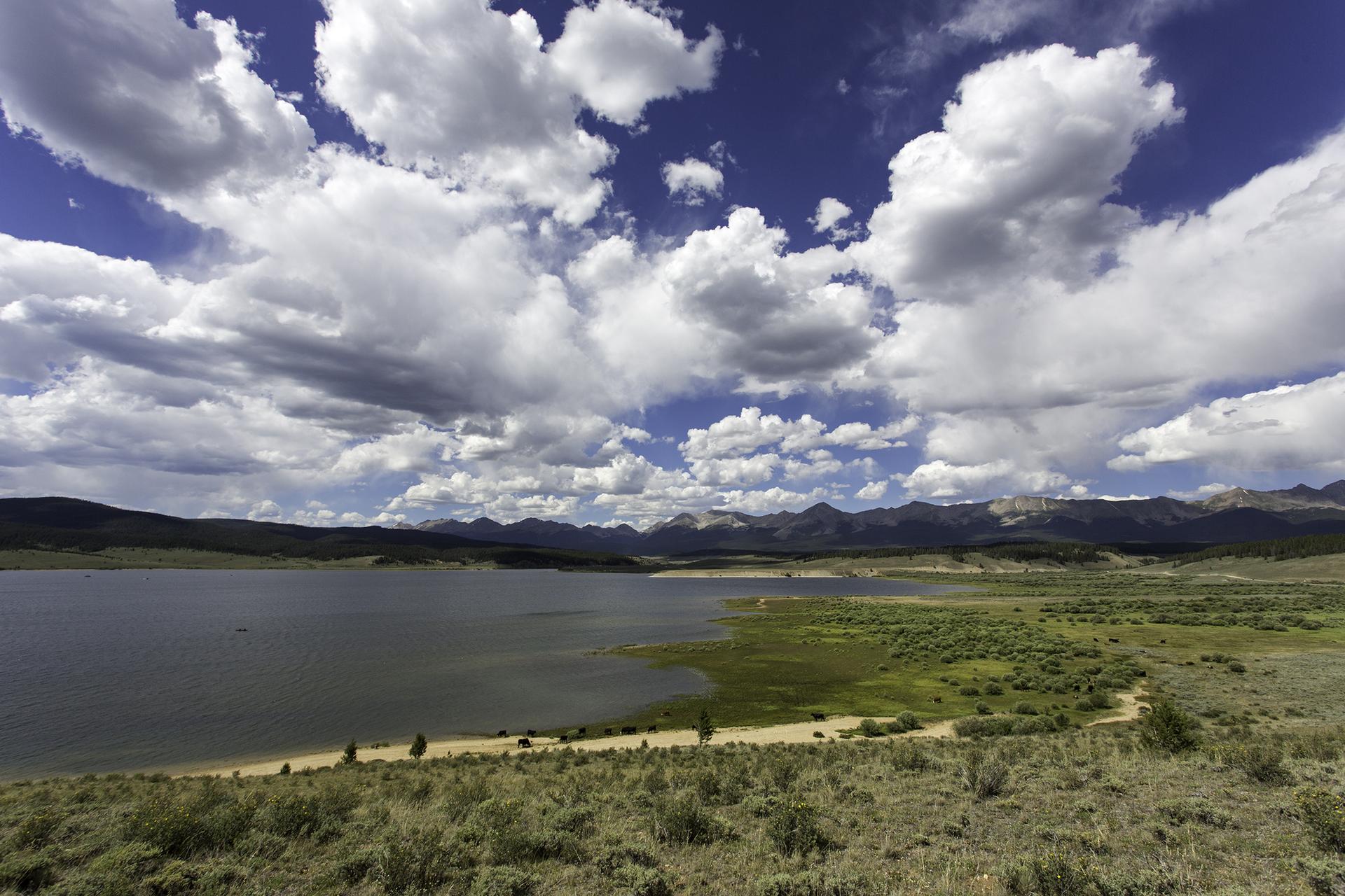 Expansive lake with green shores, distant mountains, and dramatic cloudy sky.