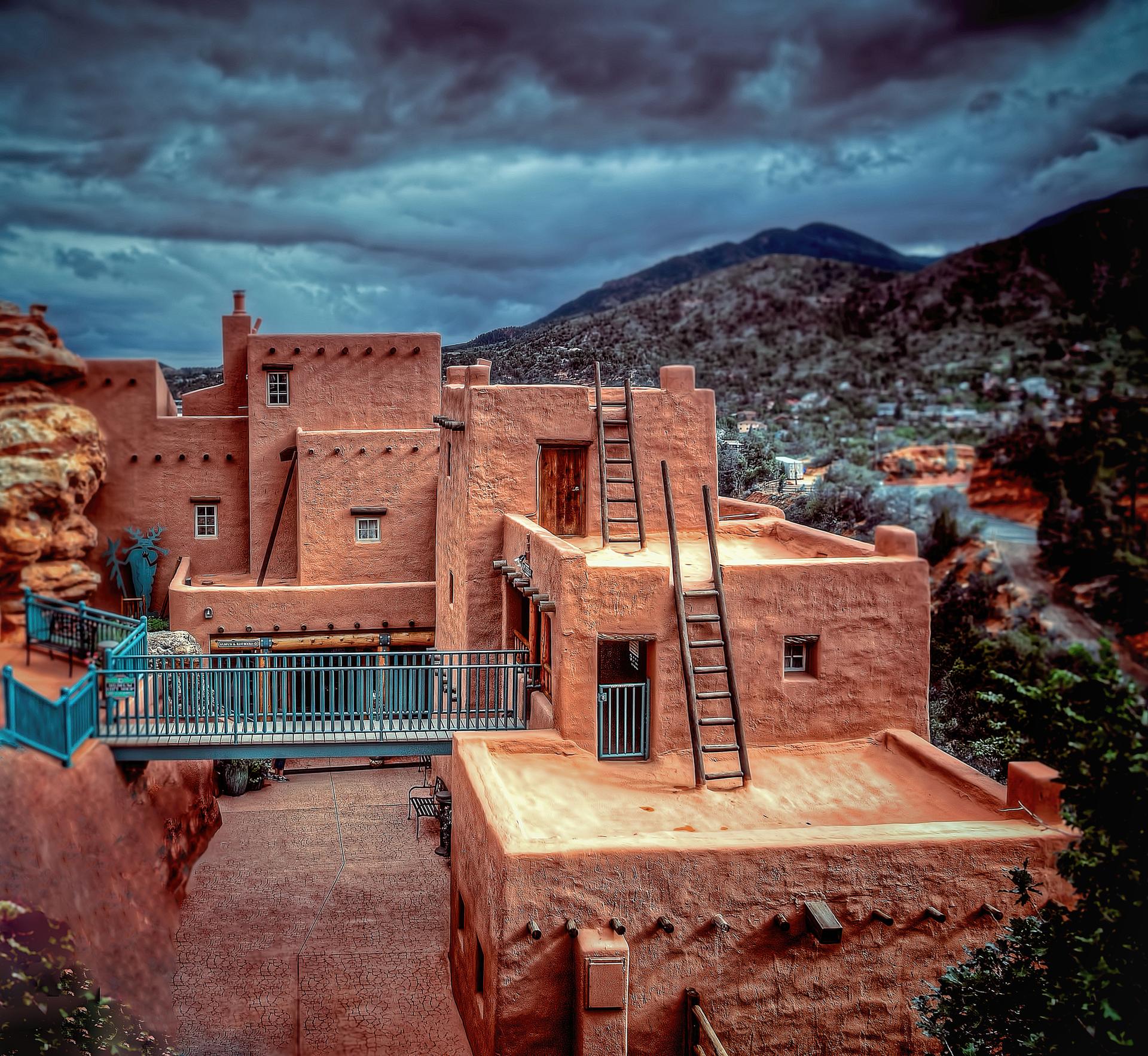 Terracotta buildings with ladders and a blue bridge under dark, cloudy skies, mountains in background.