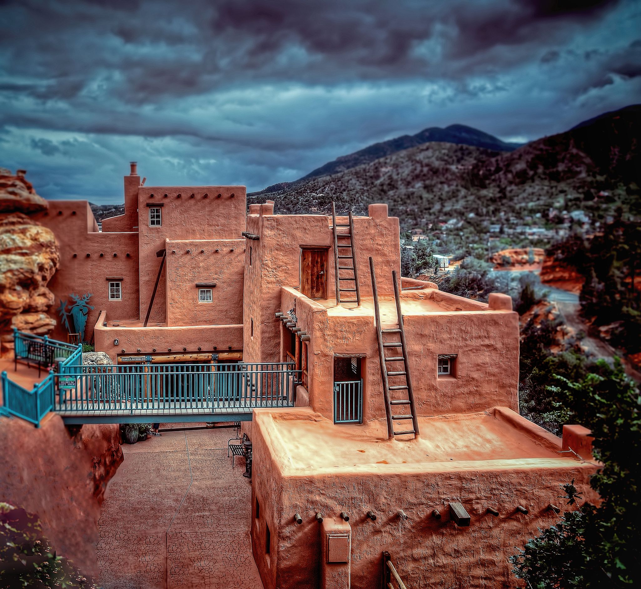 Terracotta buildings with ladders and a blue bridge under dark, cloudy skies, mountains in background. - Manitou Cliff Dwellings loop