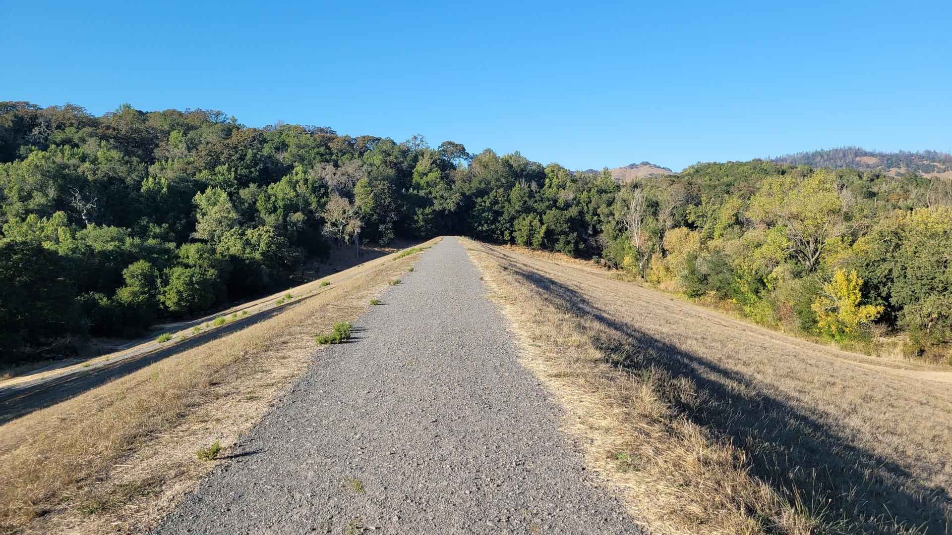 A gravel path stretches between green trees and dry grass under a clear sky.