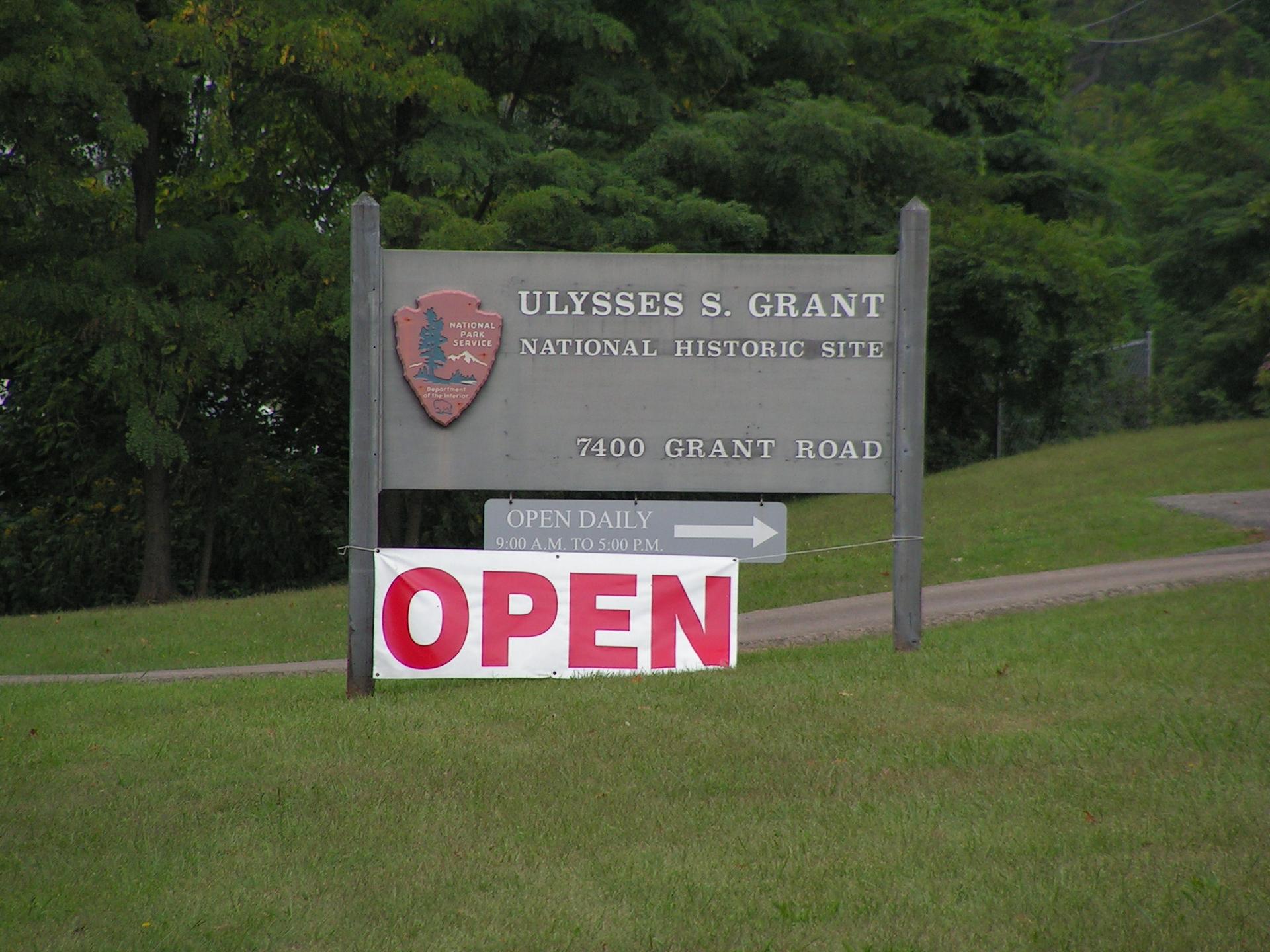 Wooden sign with "OPEN" banner on a grassy hill with trees.