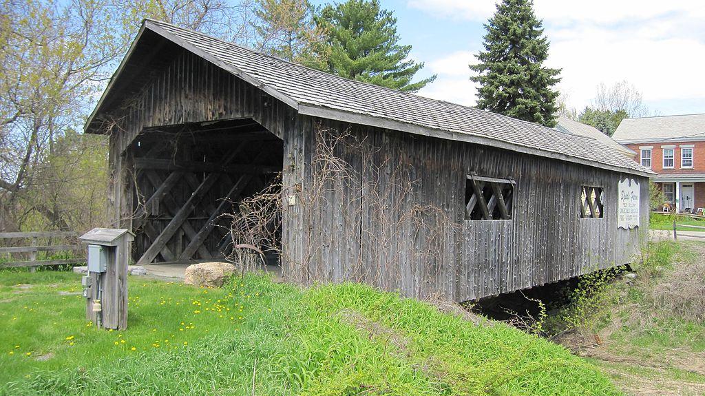 Weathered wooden covered bridge with trees and a building under bright skies.