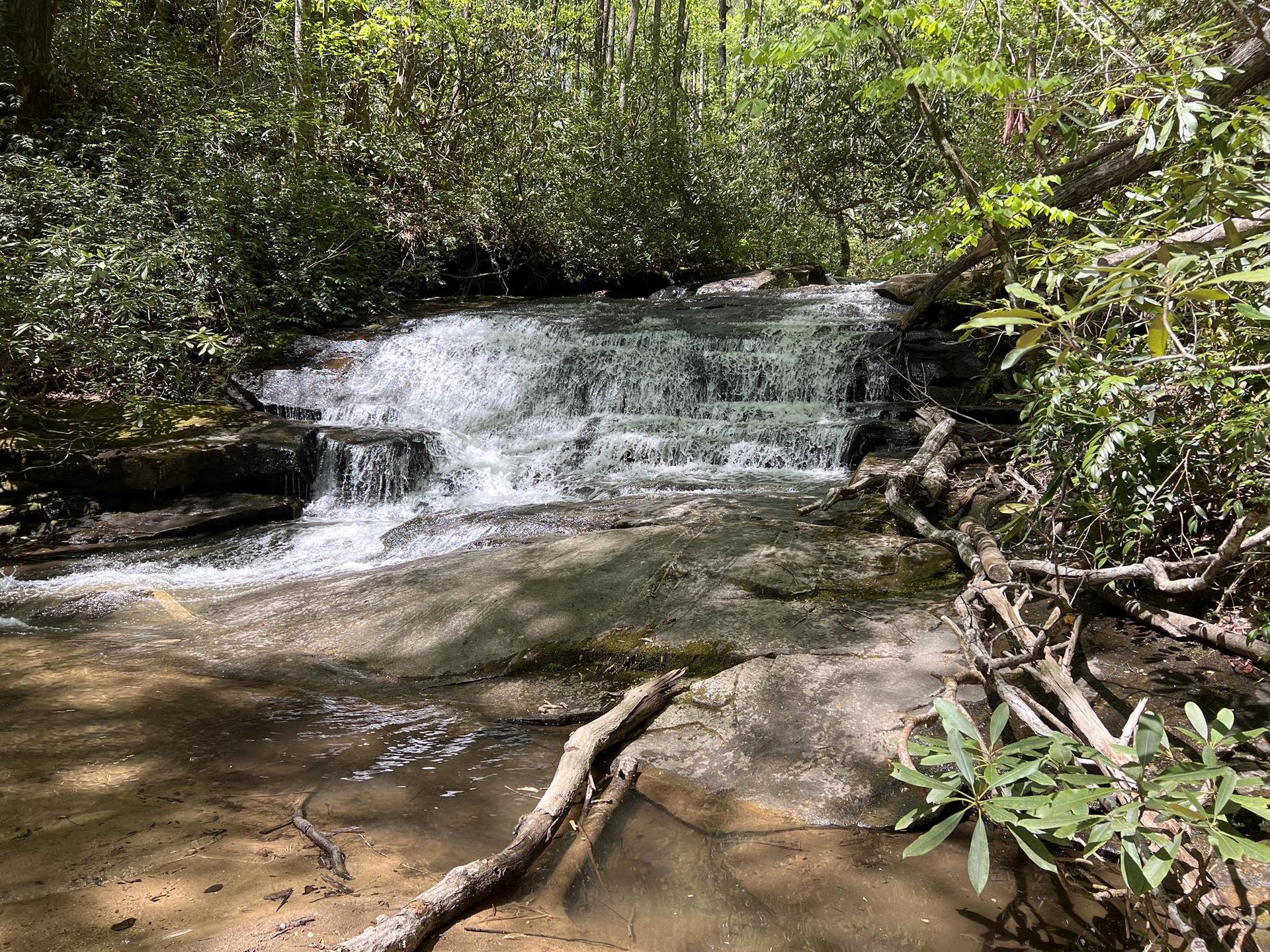 Wide, tiered waterfall flows over rocks in a lush green forest with dappled light.