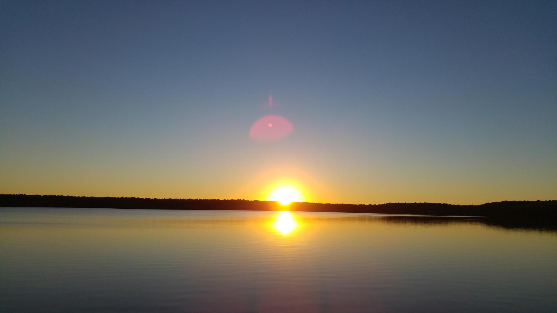 Bright sunset over a calm lake, with sun reflecting on the water.
