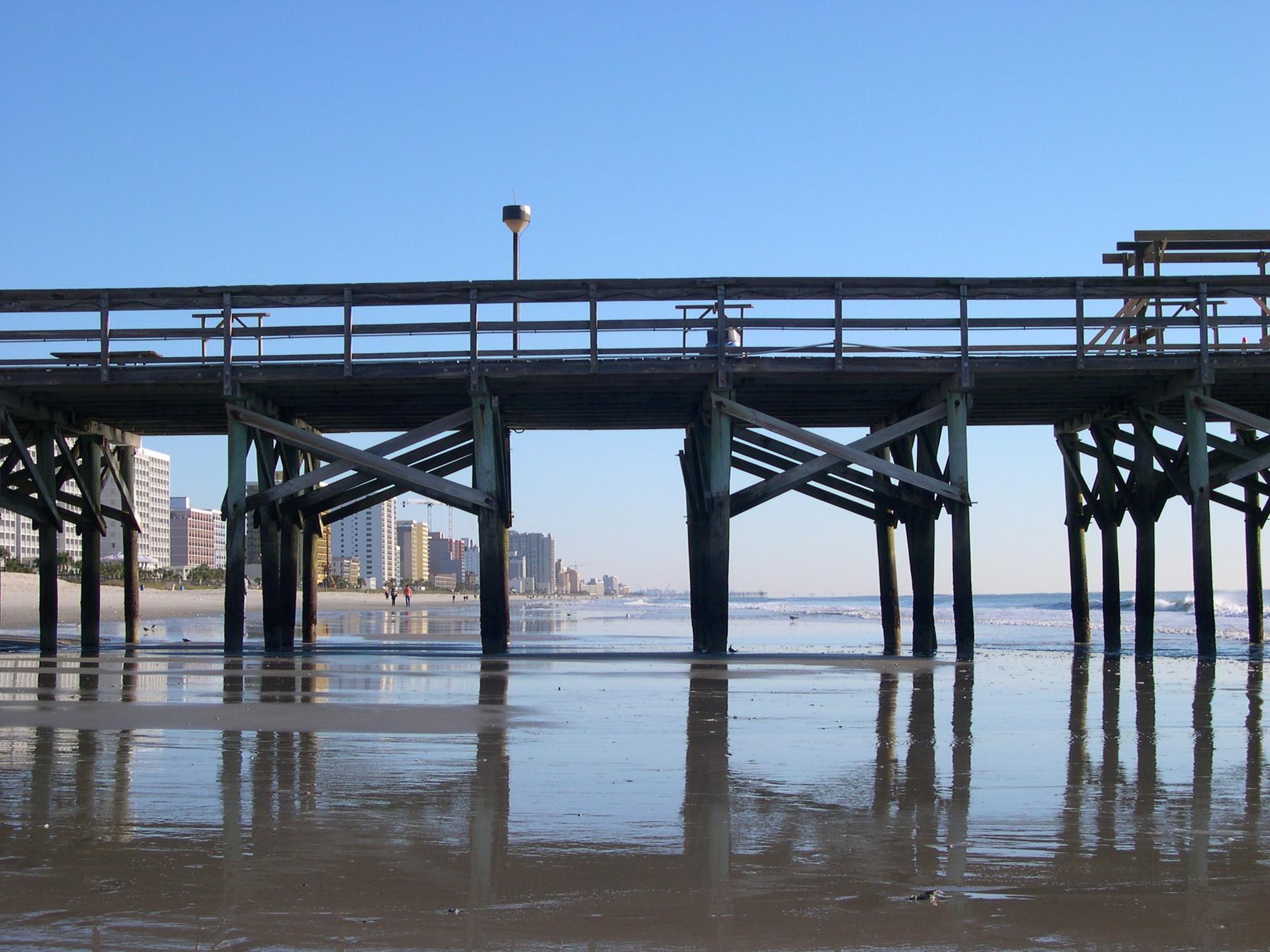 Wooden pier over calm water on a sunny beach, with a city in the background.