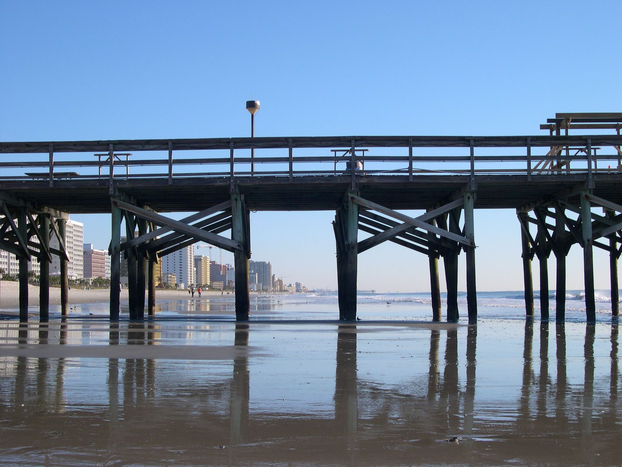 Wooden pier over calm water on a sunny beach, with a city in the background. - Springmaid Pier loop from Myrtle Beach State Park