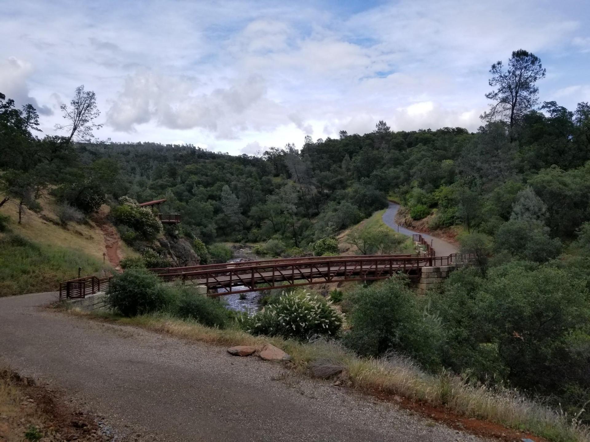 Gravel path leading to a metal bridge over a river in a green, cloudy landscape.