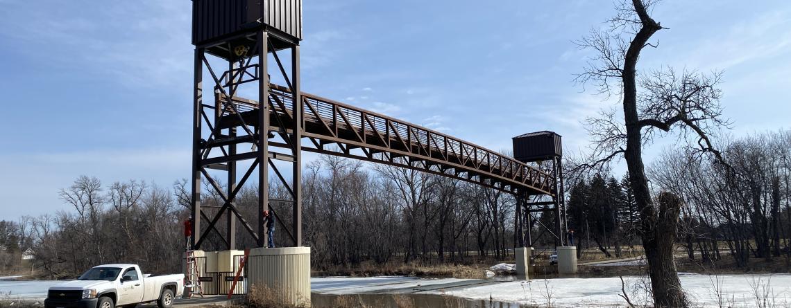 Tall metal pedestrian bridge over icy ground with bare trees, bright day.