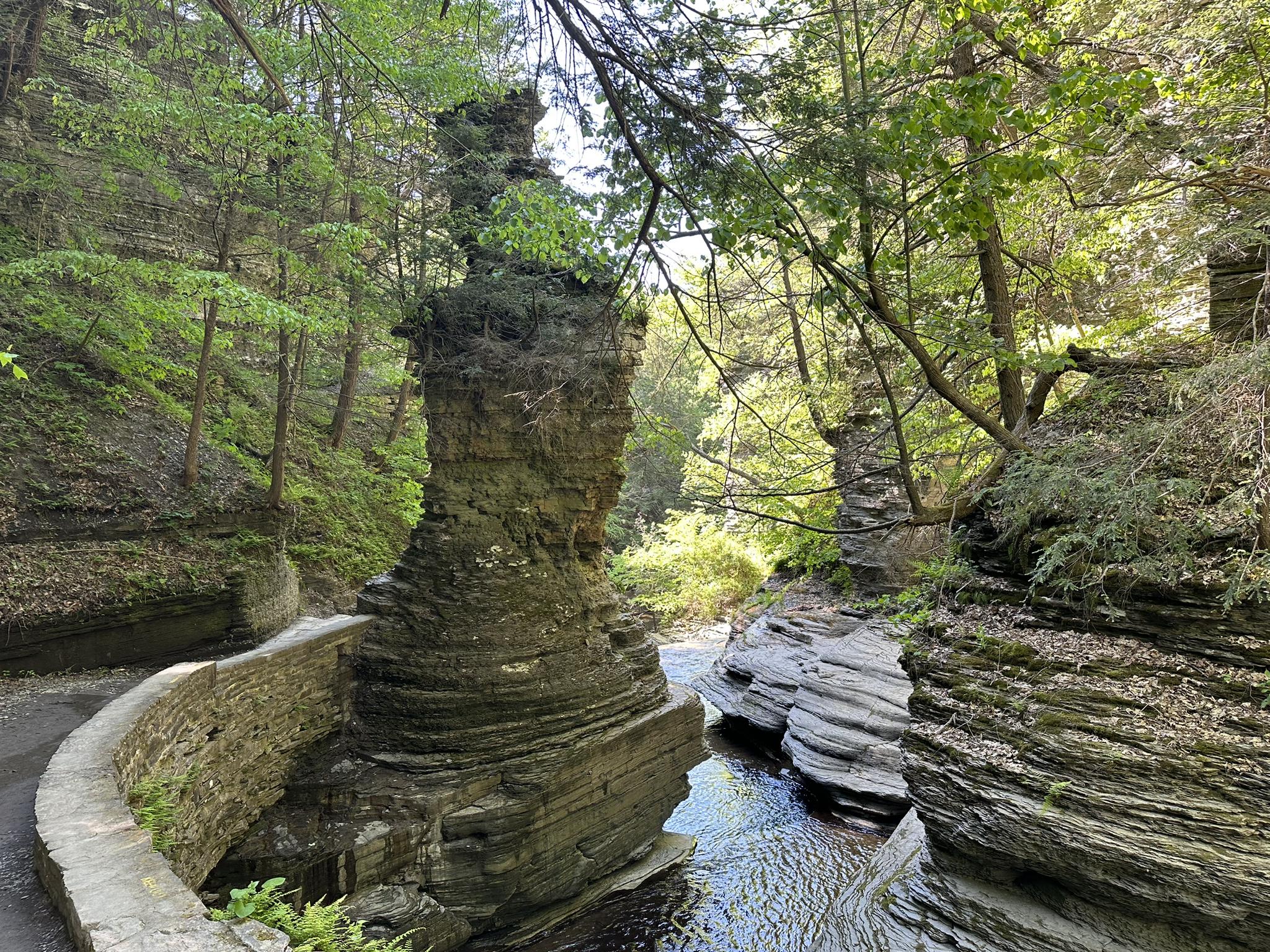 Sunlit gorge with unique rock formations, flowing water, and a stone path.