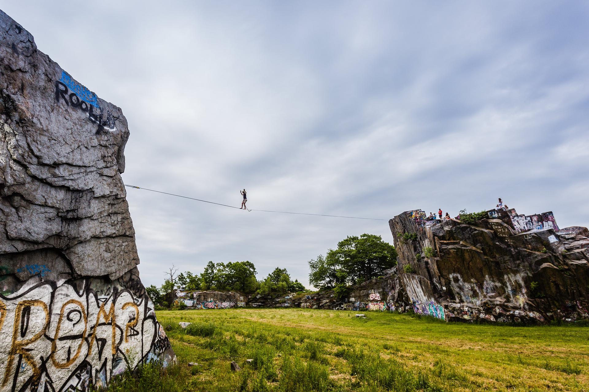 Person slacklining between graffiti-covered rocks over a grassy field under cloudy sky.