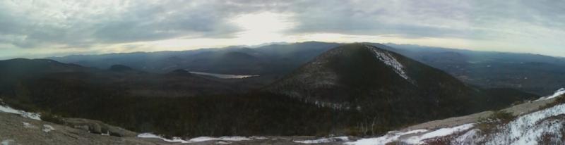 Panoramic view of forested mountains, distant lake, and patches of snow.