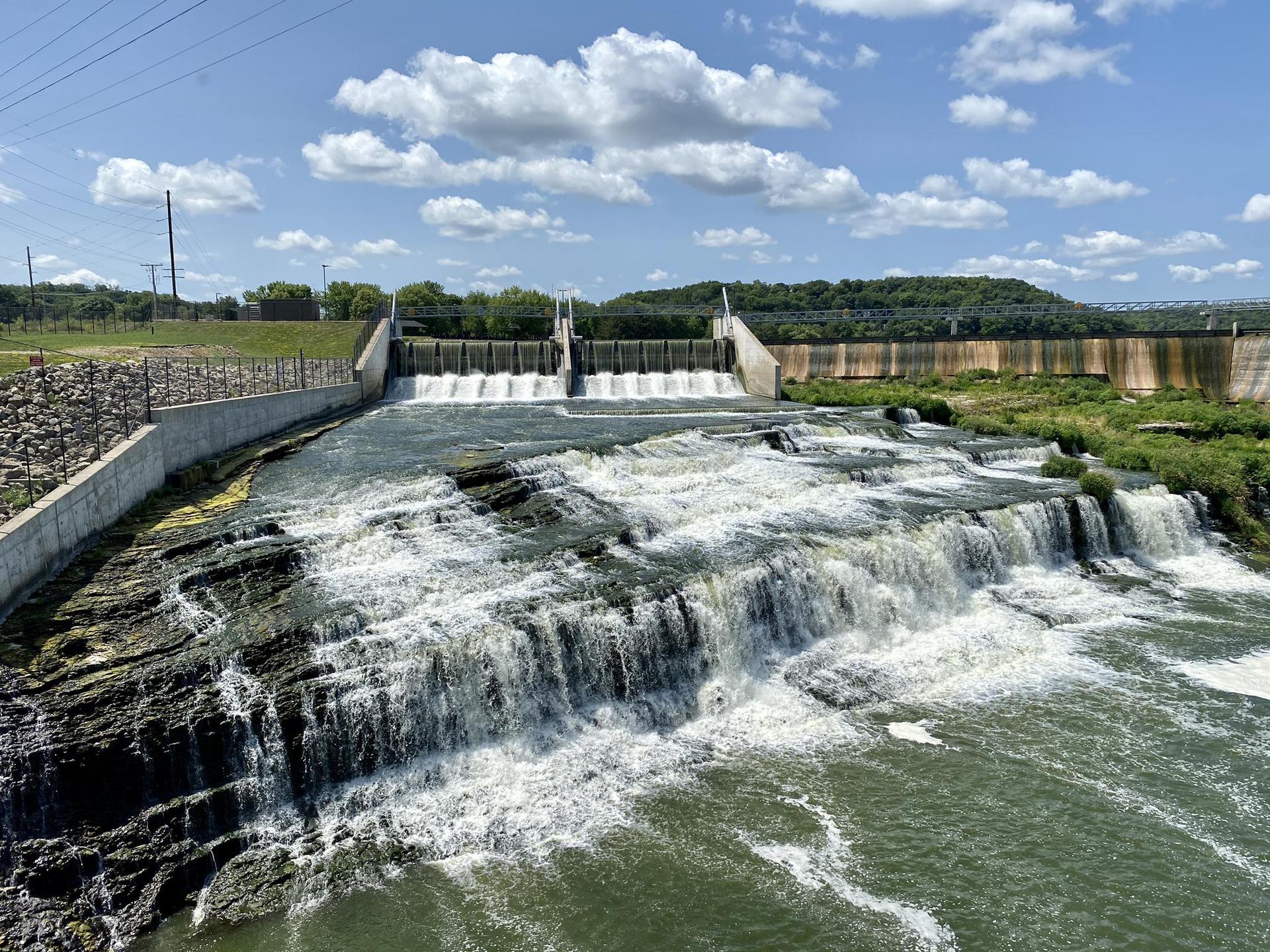 Dam with water cascading over rocks under a bright, cloudy sky.