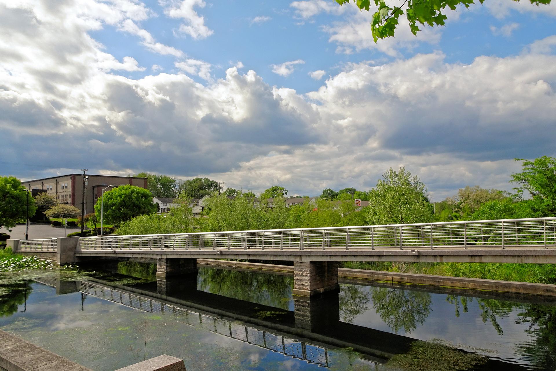 A pedestrian bridge spans calm water under a partly cloudy sky, surrounded by green trees.