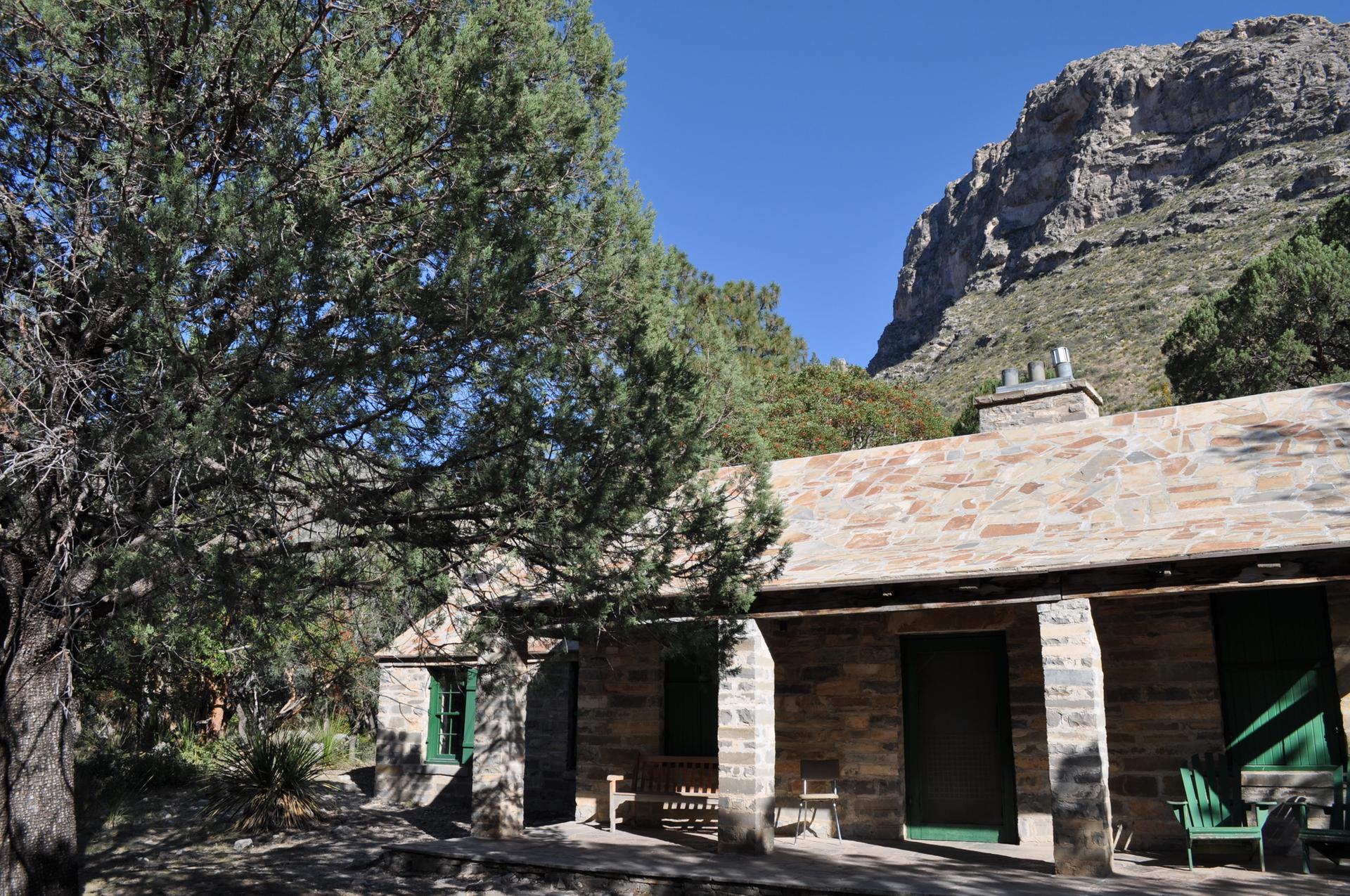 A stone building with a porch sits among trees, backed by a large mountain.