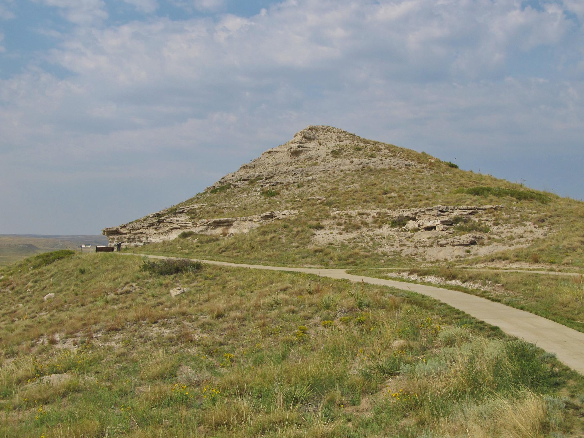 A winding road ascends a grassy, conical hill under a bright, cloudy sky.