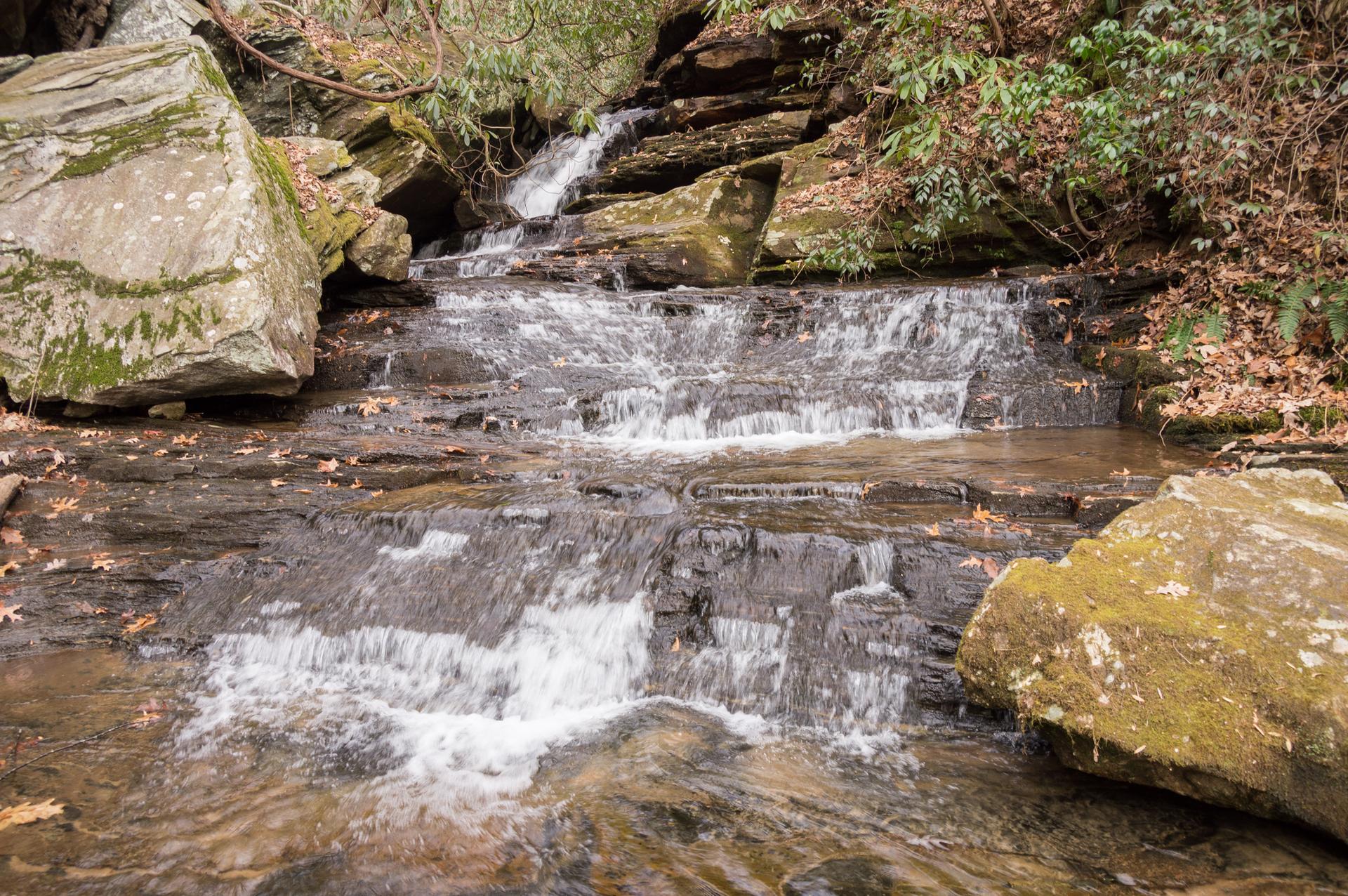 Small, multi-tiered waterfall flows over dark rocks in a wooded stream.