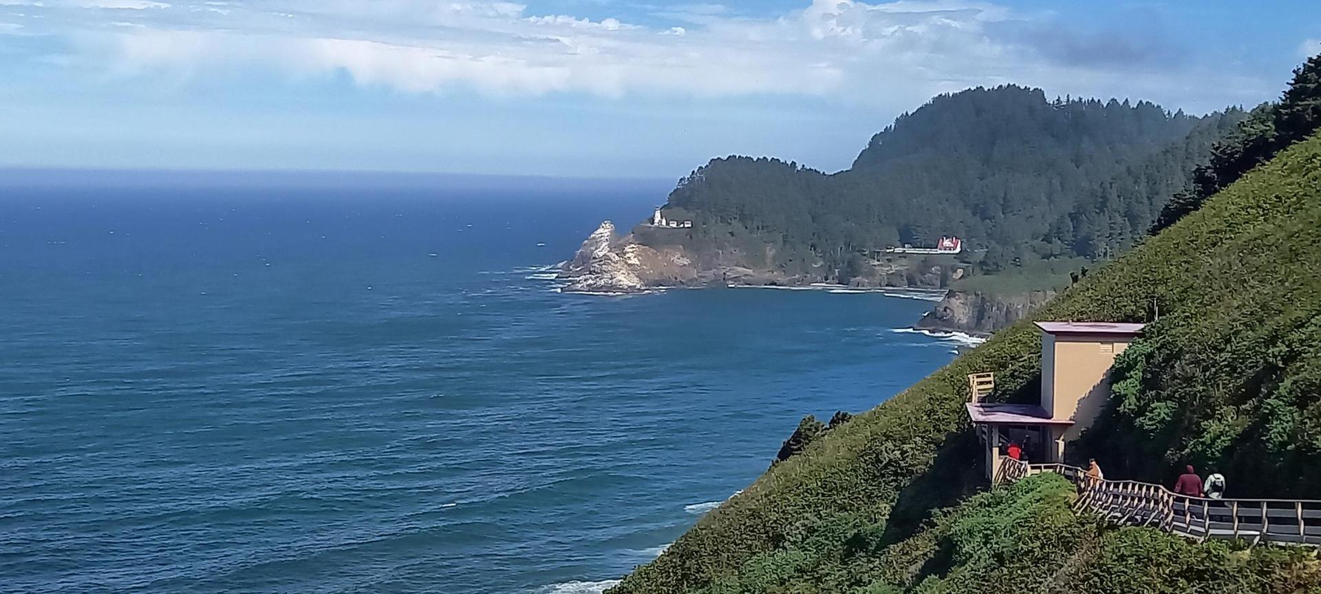 Sunny coastal view with blue ocean, distant lighthouse, and cliffside building.