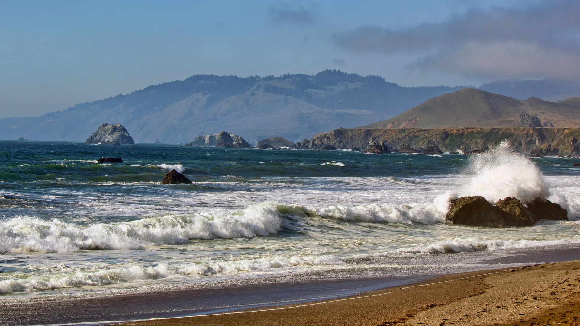 Powerful ocean waves crash against a large rock on a sandy beach.