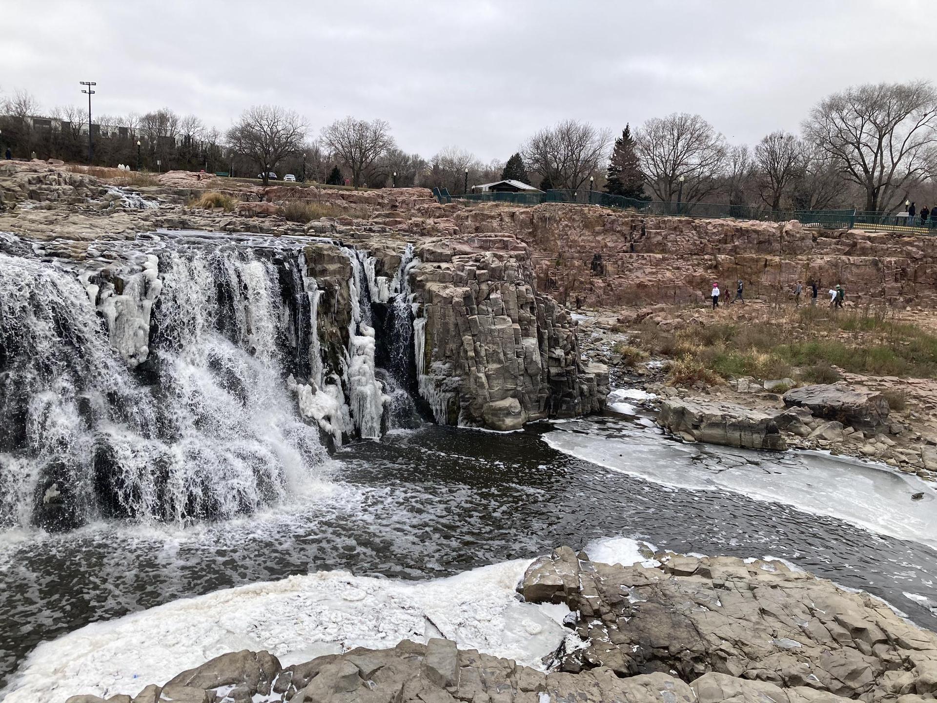 Winter scene of a partially frozen waterfall and icy river under an overcast sky.