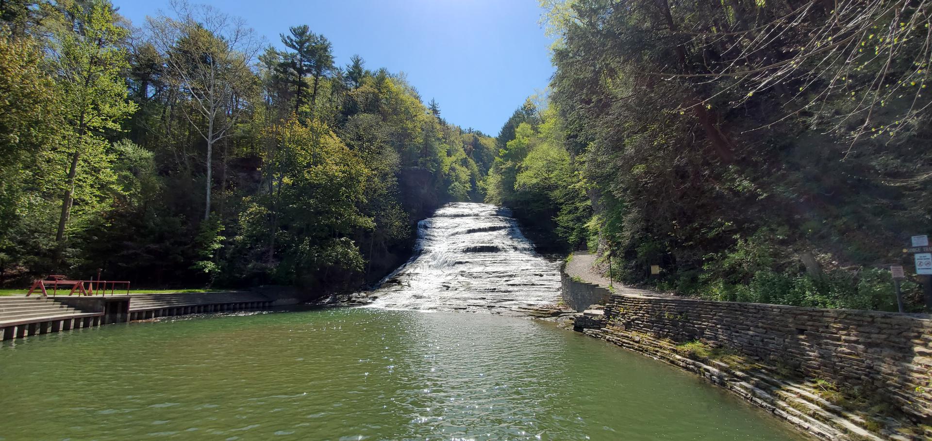 Wide waterfall flows into a green pool, surrounded by lush green forest.