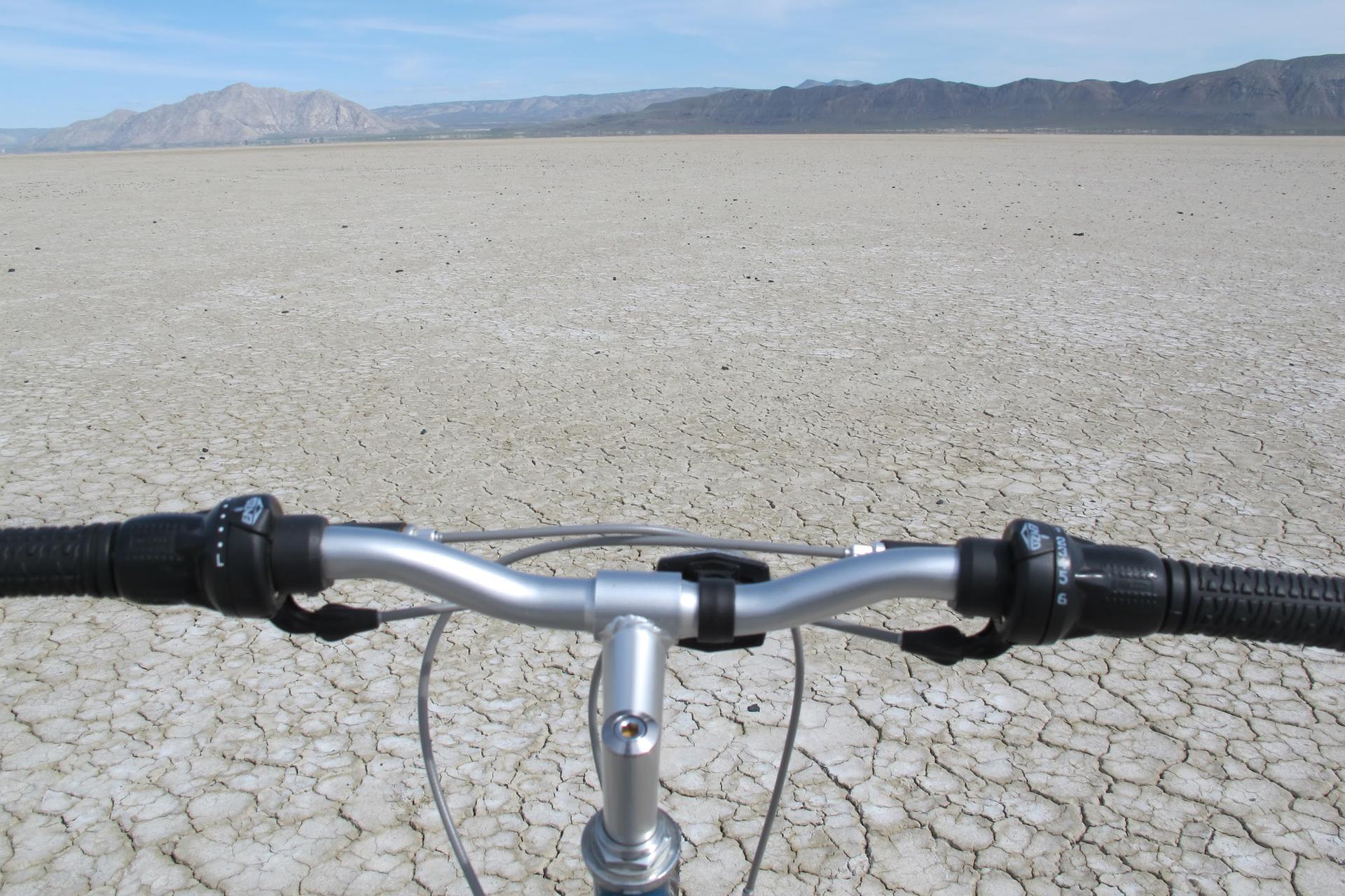Bicycle handlebars over a vast cracked dry lakebed under a bright sky.