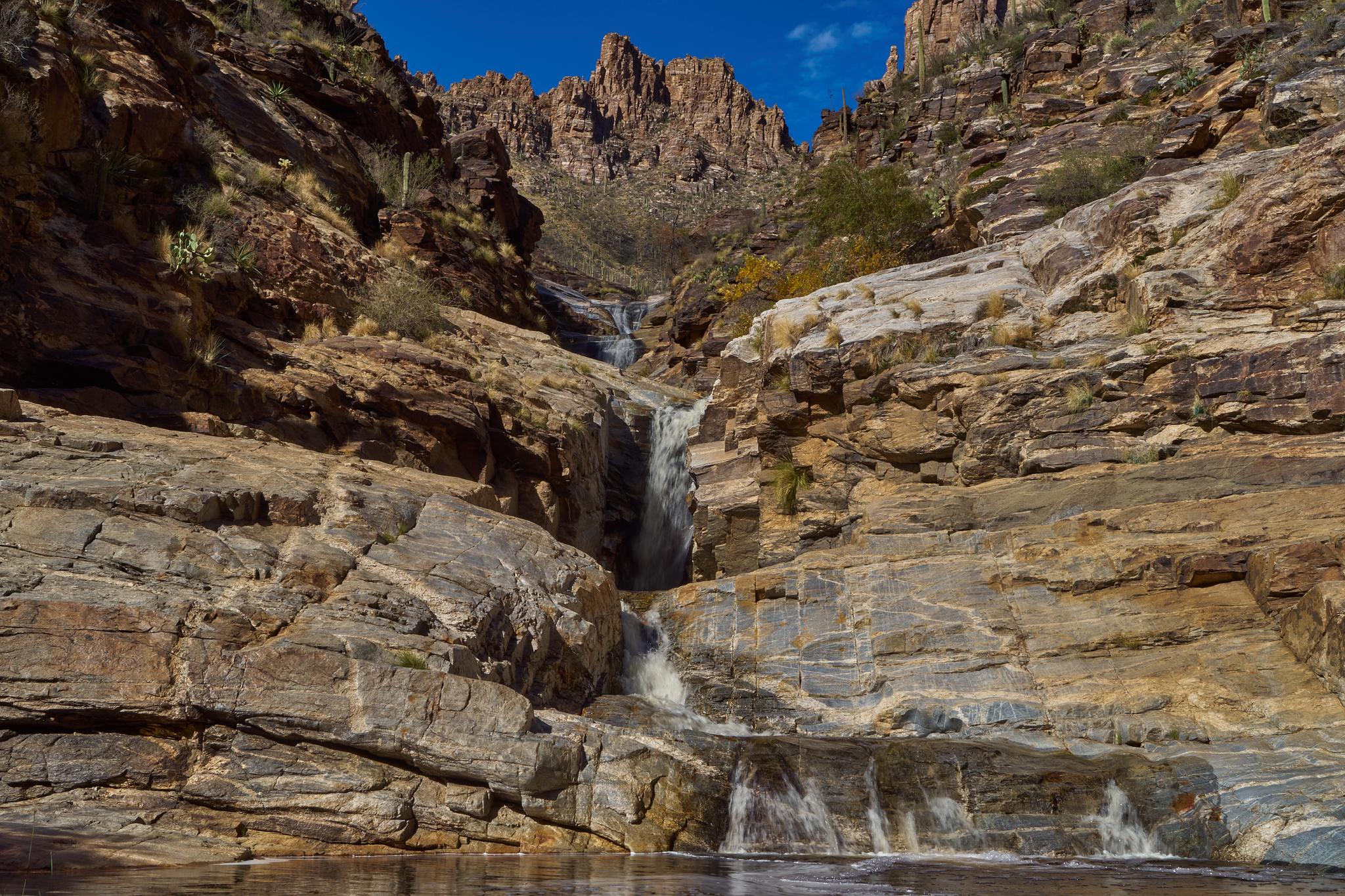 Waterfall cascades down rocky canyon walls into a clear pool under a bright blue sky.