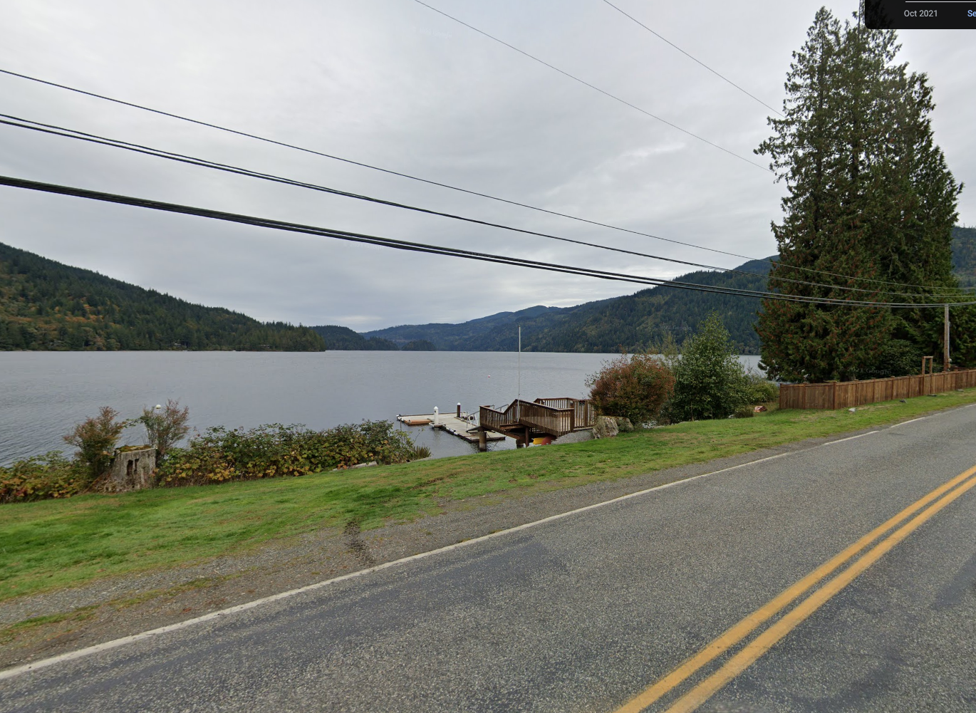 Roadside view of a calm lake, forested hills, and a dock with small boats.