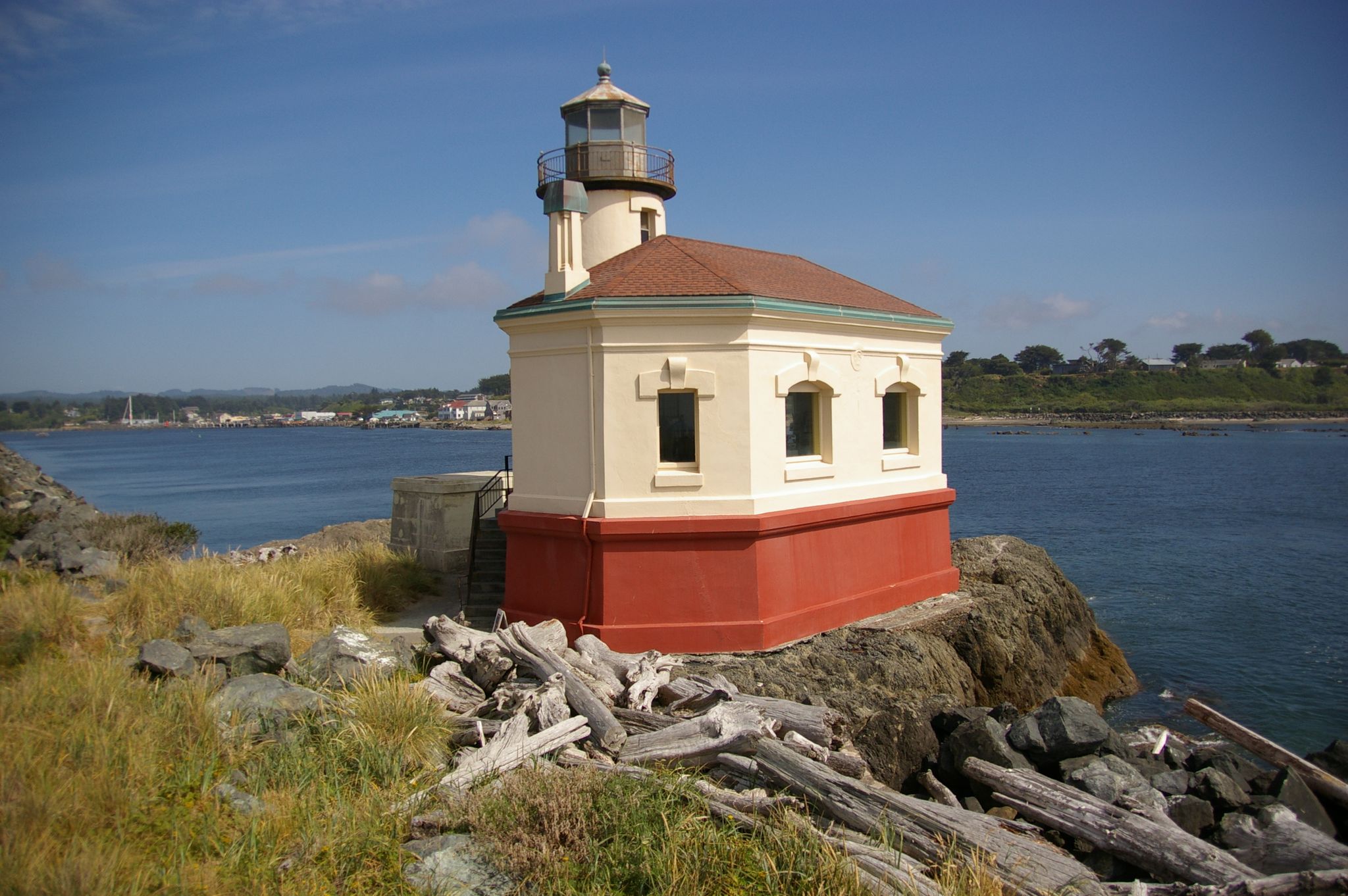 A small, cream and red lighthouse on a rocky, grassy shore under a clear sky. - Coquille River Lighthouse – View of the Coquille River loop from Bullards Beach State Park