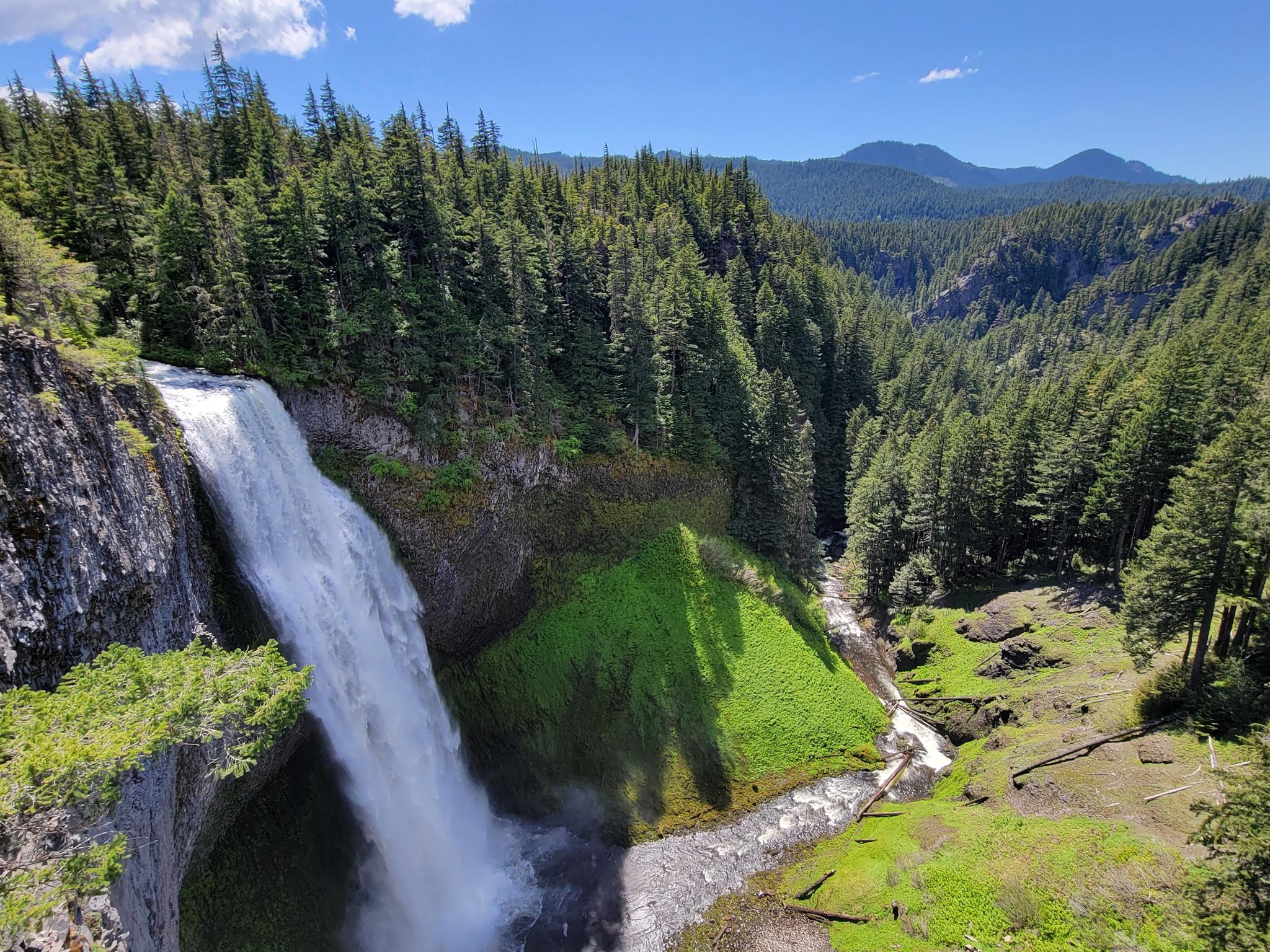 A powerful waterfall cascades into a lush green valley surrounded by forests.