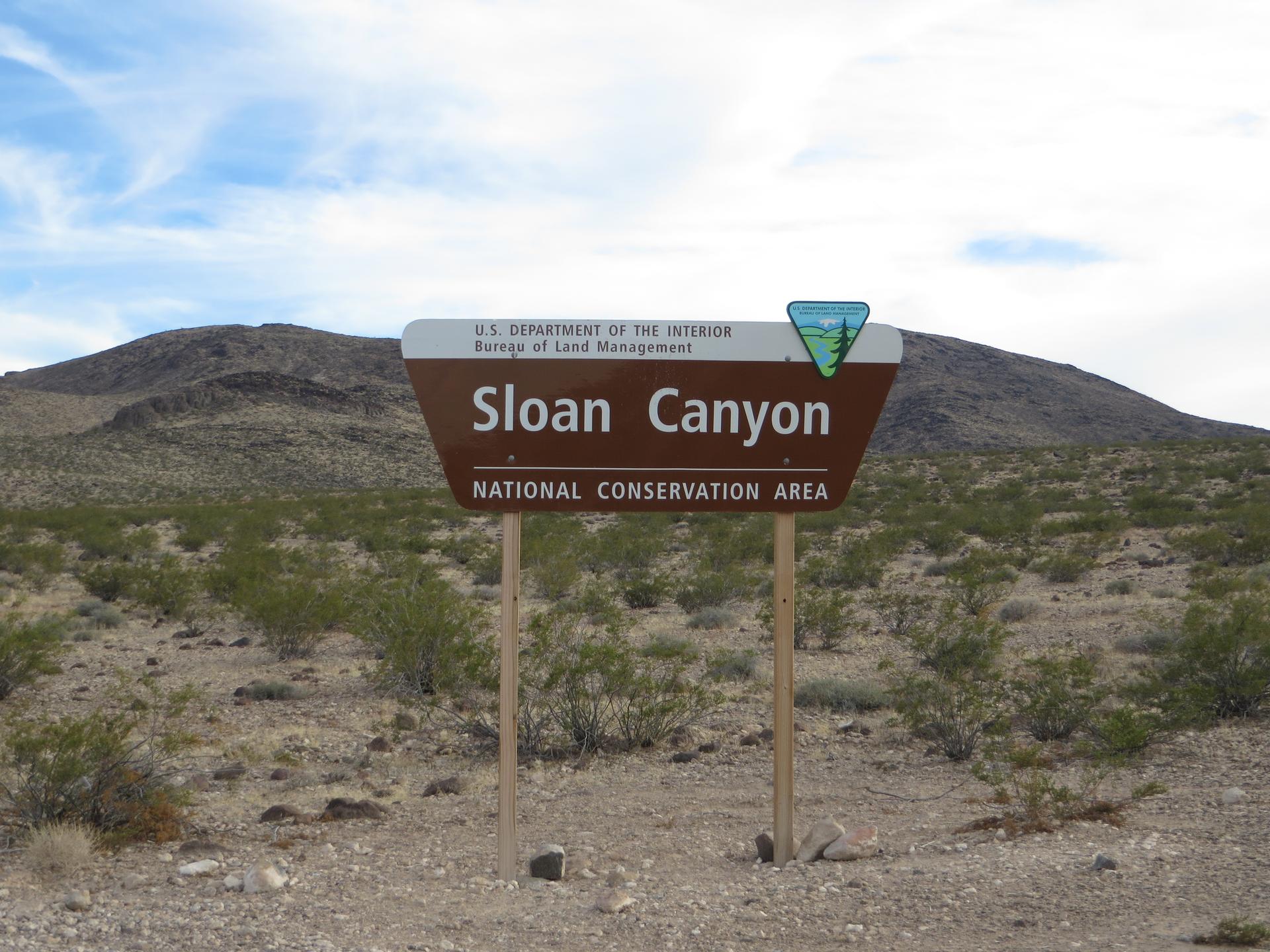 An informational sign stands in a desert landscape with hills under a partly cloudy sky.