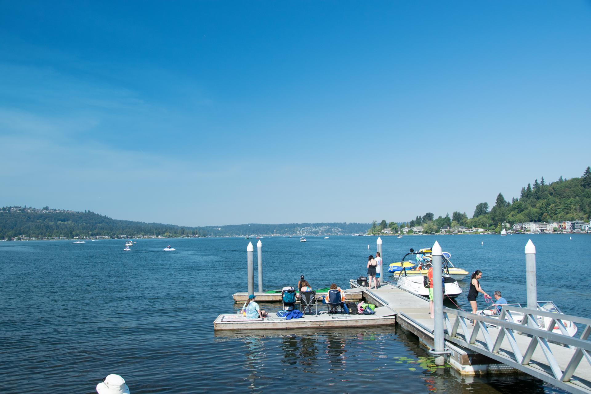 People relax on a sunny dock with boats on a wide, blue lake.