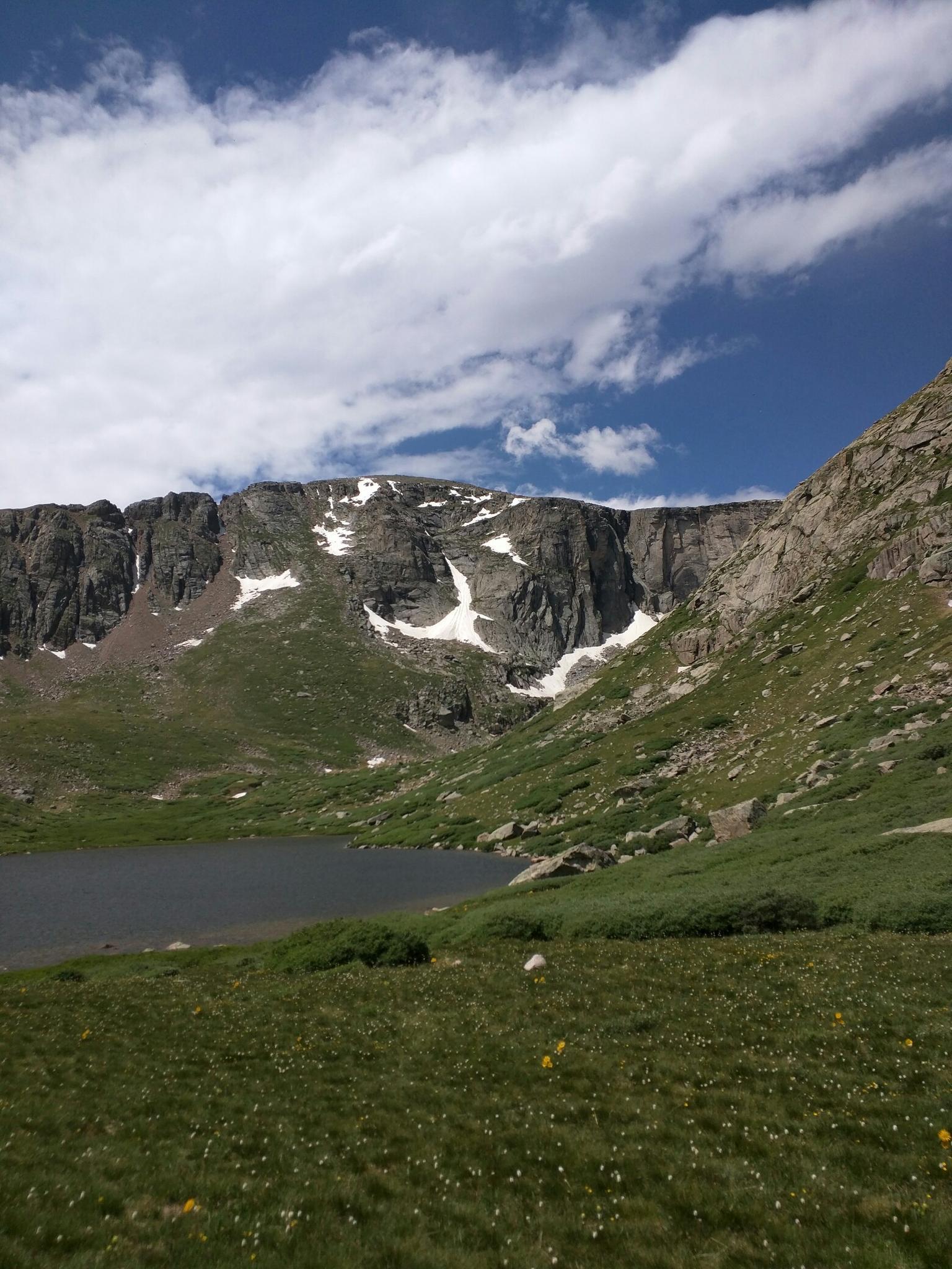 Dark mountain lake at the base of a massive rocky cliff with snow patches.