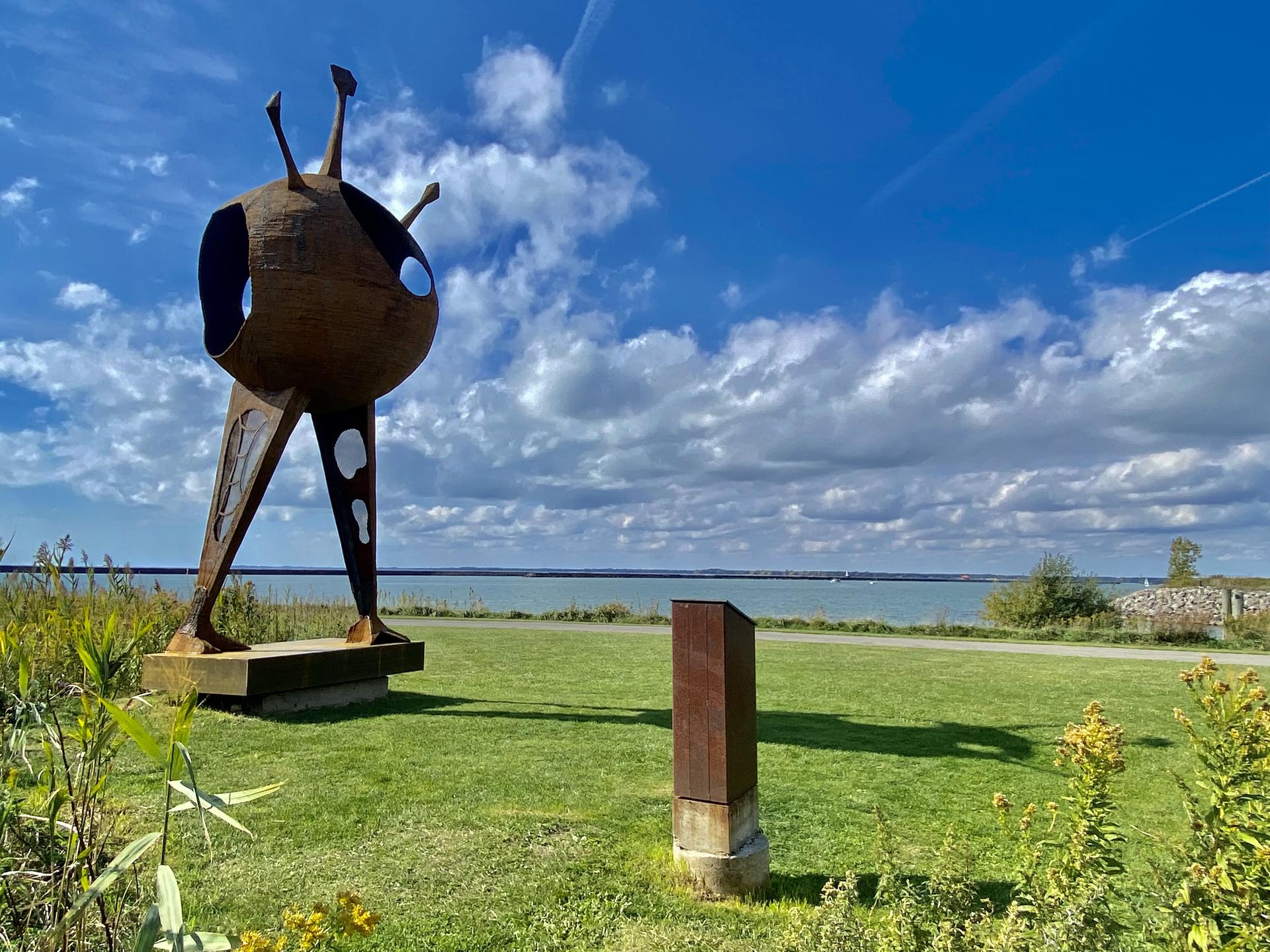 Abstract rusty metal sculpture on grass by a large body of water under a blue sky.