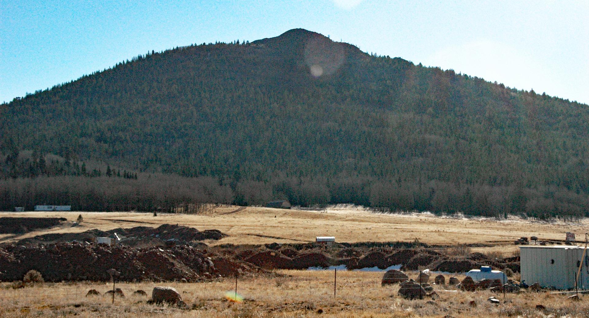 Forested mountain overlooks a sunny, dry field with earthworks and distant buildings.