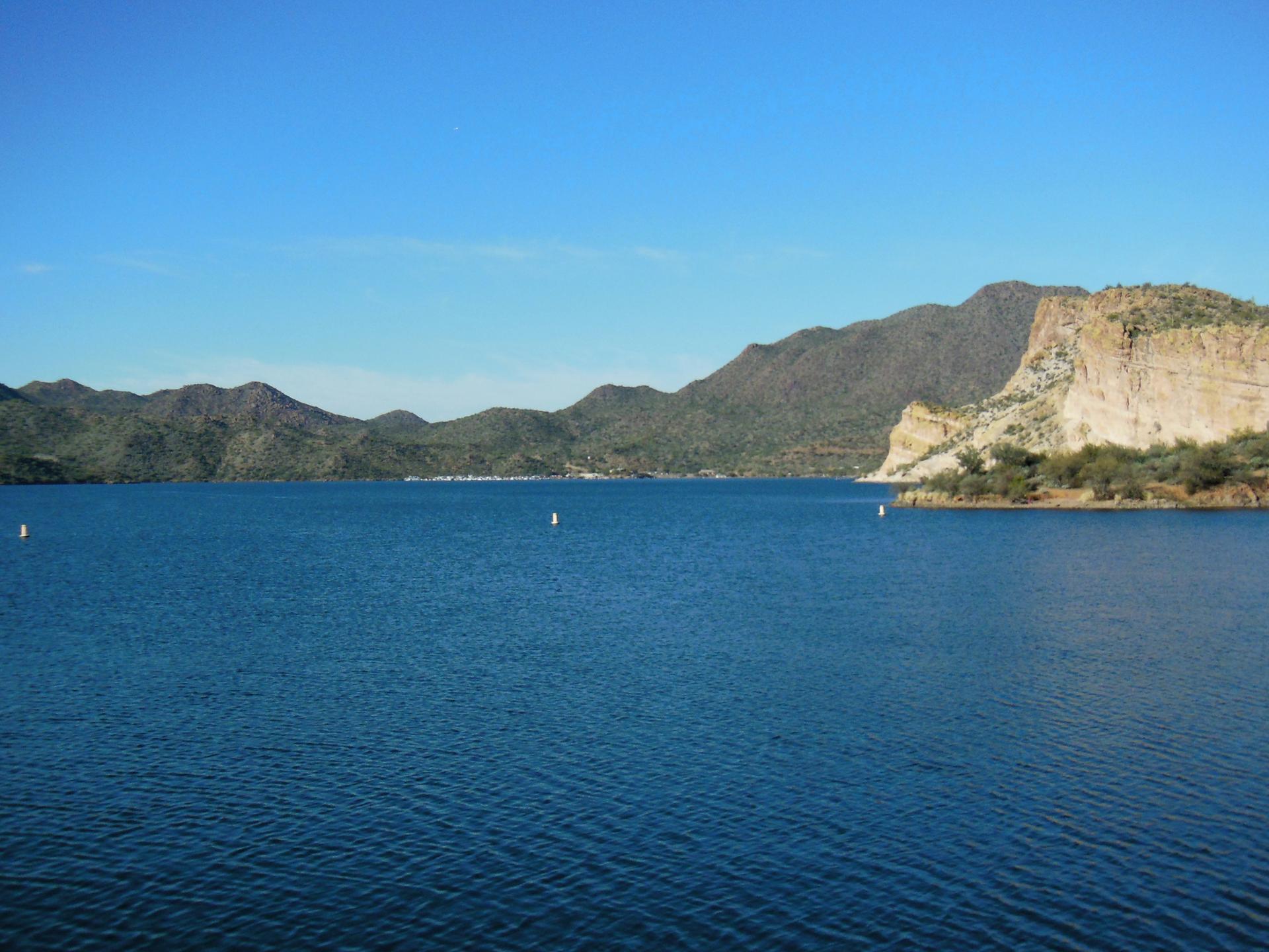 Expansive blue lake surrounded by green mountains and a rocky cliff in daylight.