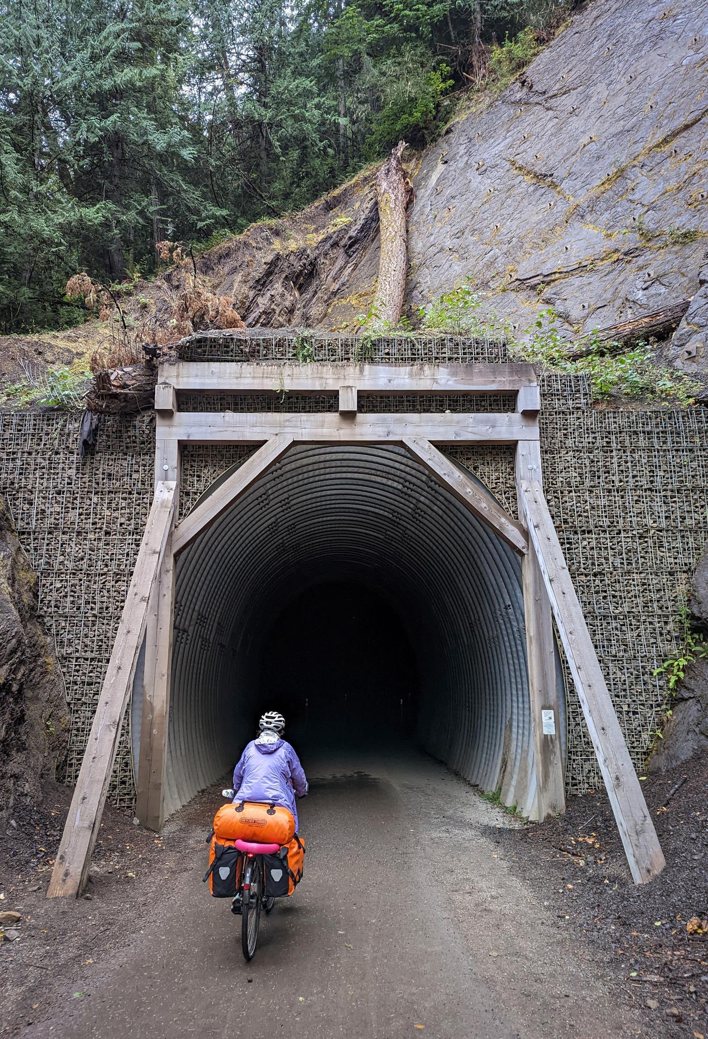 Cyclist on a dirt path entering a dark, corrugated metal tunnel.