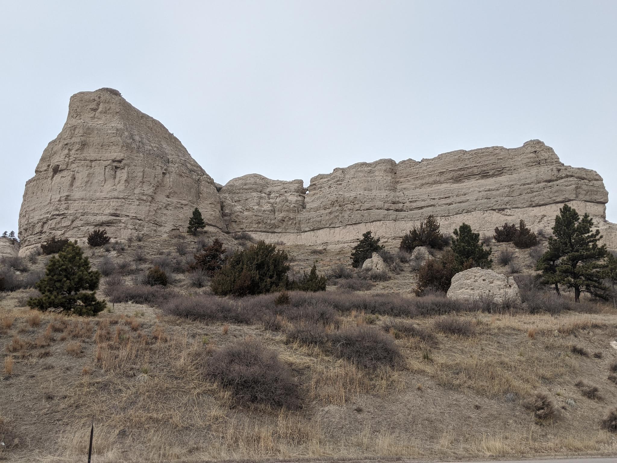 Large, light-colored rock formations rise above dry, brushy hills under an overcast sky.