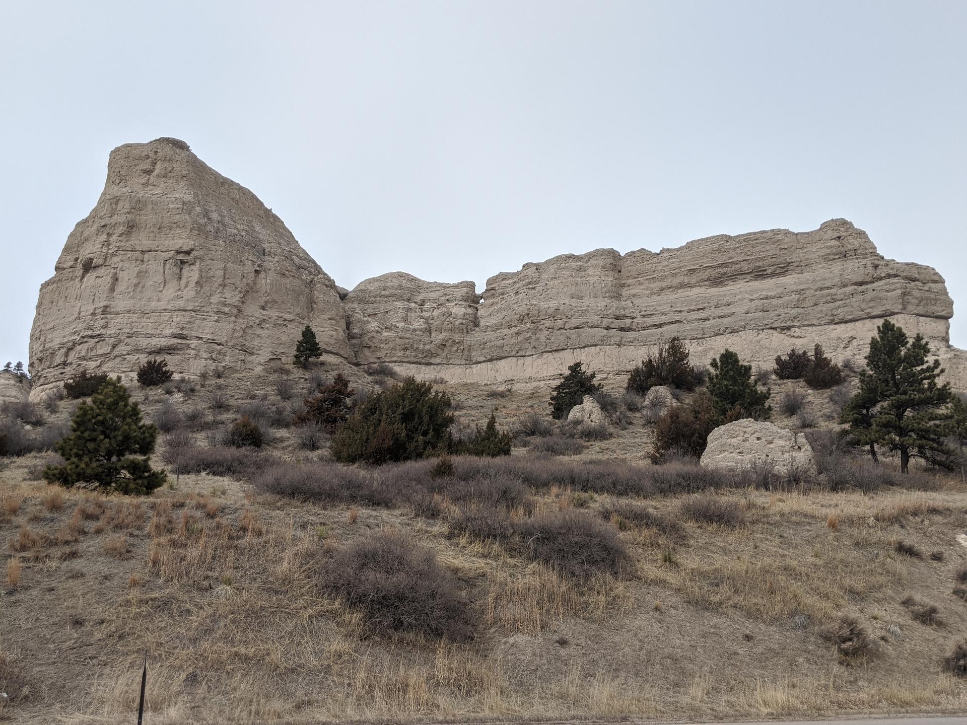 Large, light-colored rock formations rise above dry, brushy hills under an overcast sky.