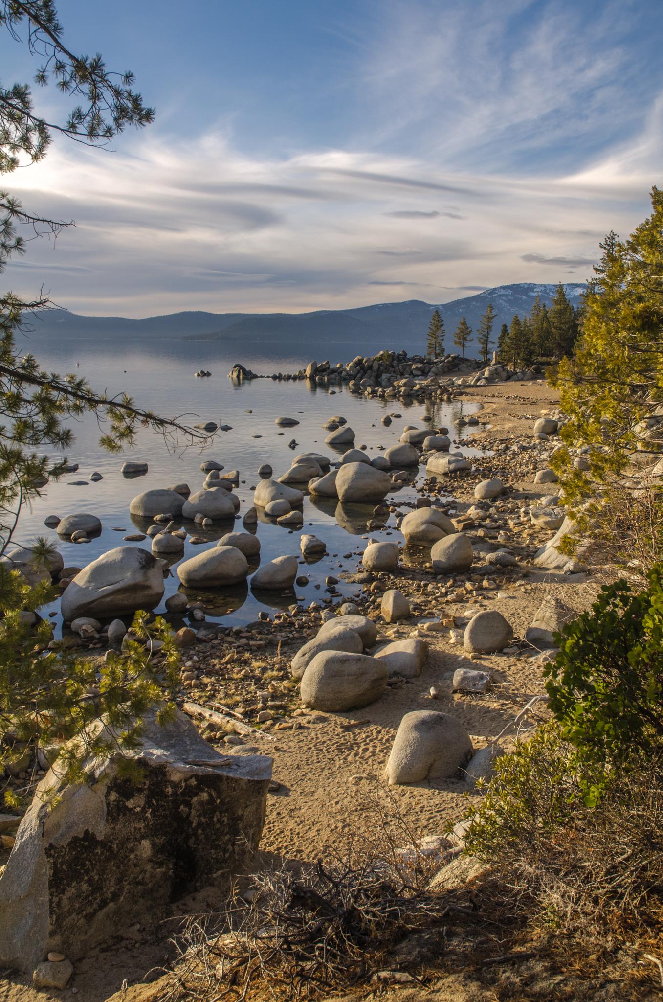 Sunny, rocky beach with calm lake water and distant mountains under a blue sky.