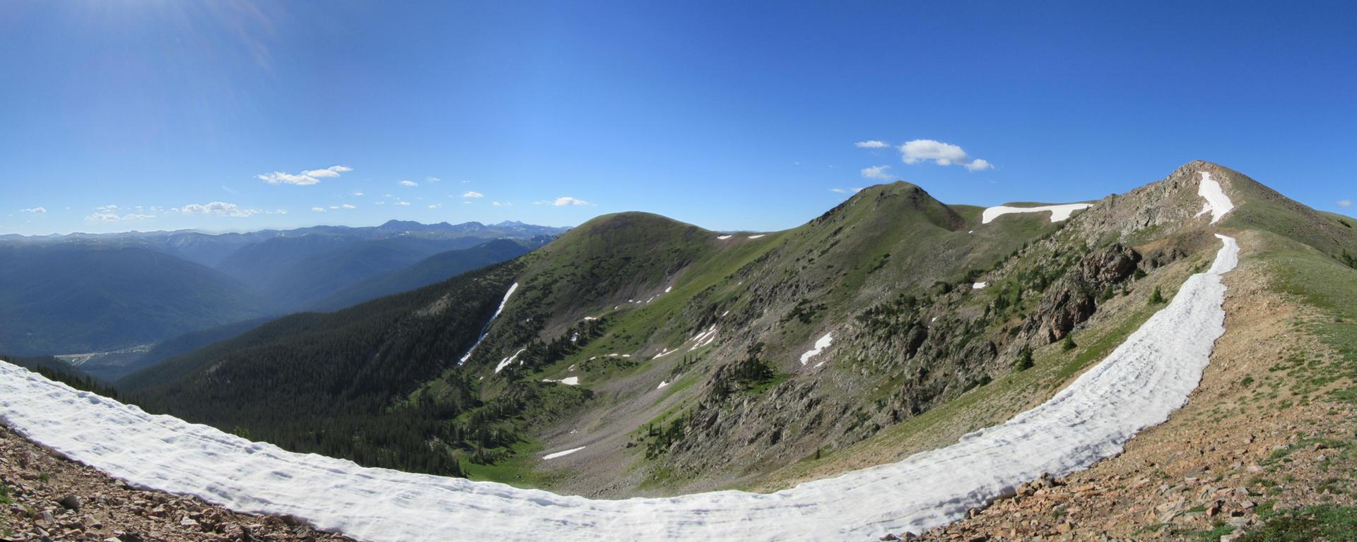 Panoramic view of rugged mountains with snow patches under a bright blue sky.