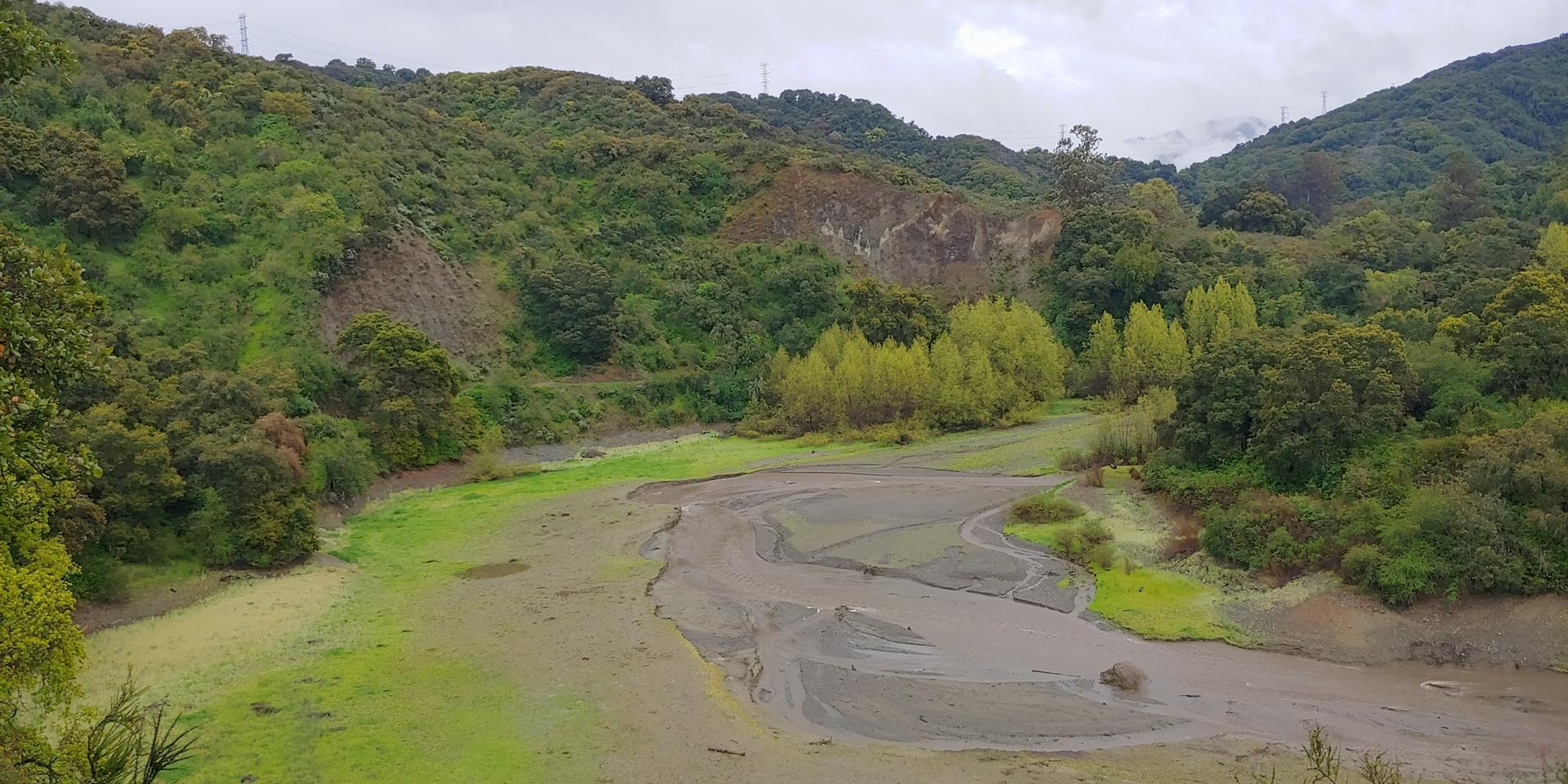 Overcast view of a muddy stream winding through a green, hilly landscape.