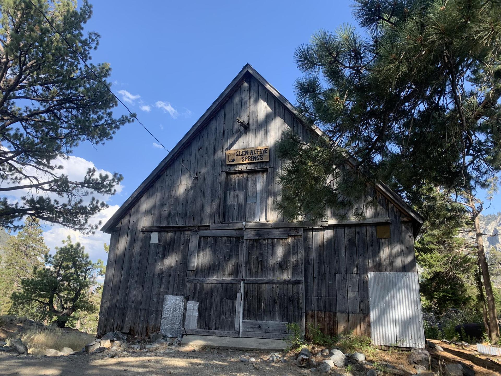 An old wooden barn-like building stands among trees on a sunny day.