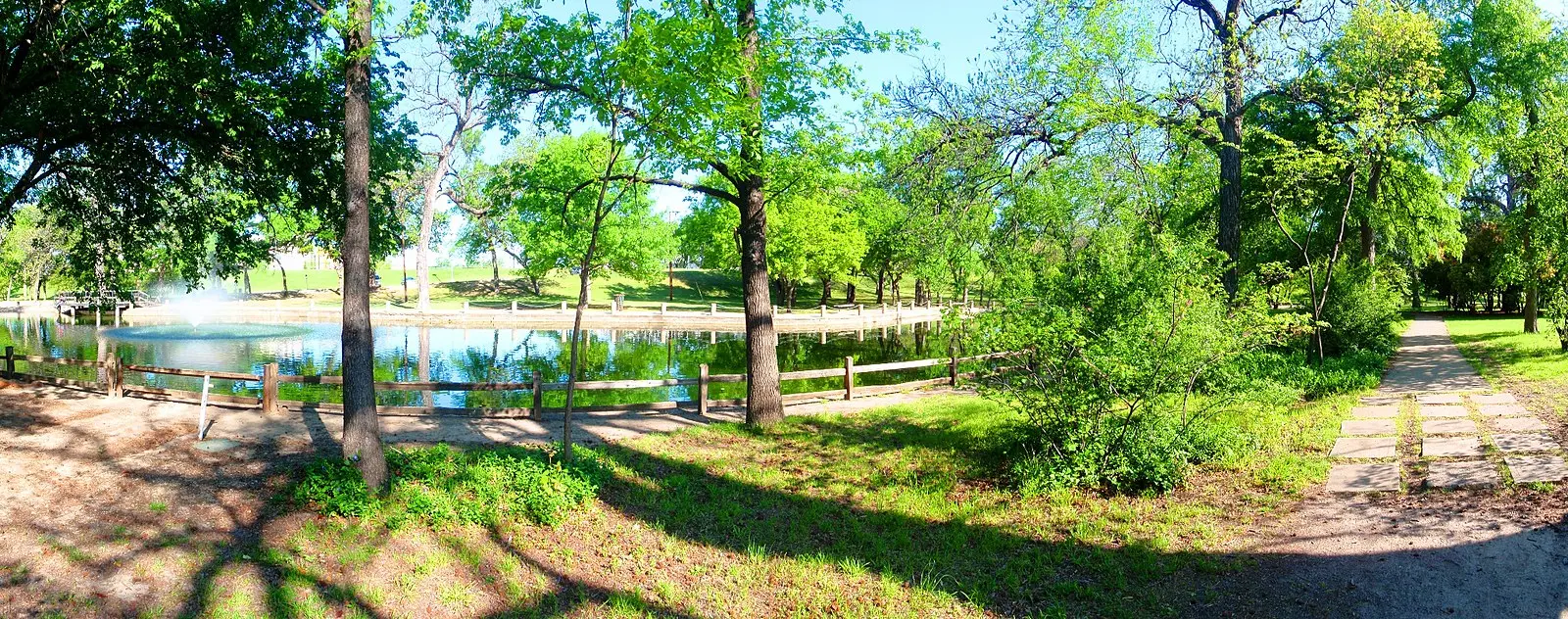 Sunny park scene with a pond, fountain, and lush green trees.