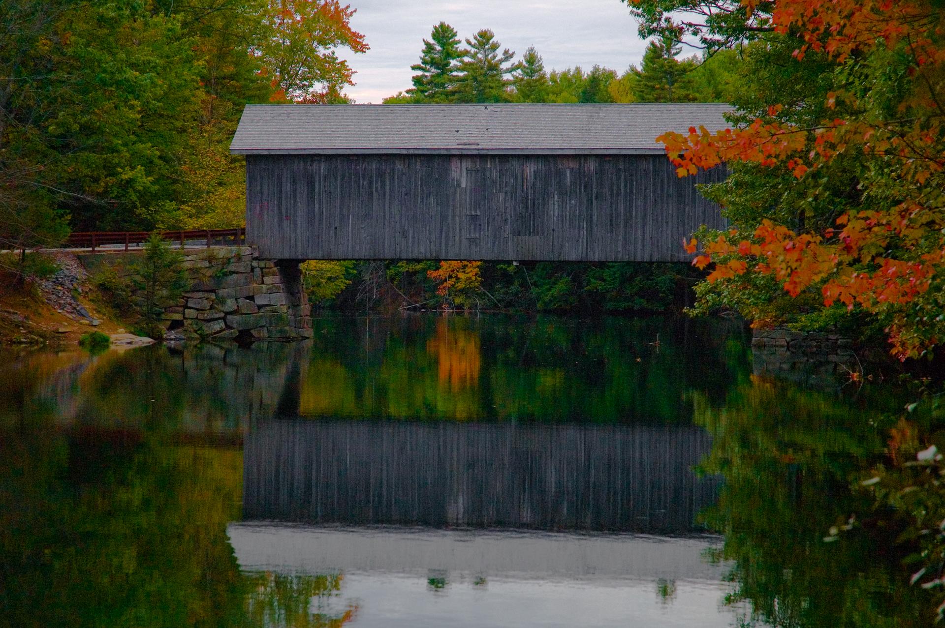 A covered wooden bridge spans calm water, surrounded by vibrant autumn foliage under an overcast sky.