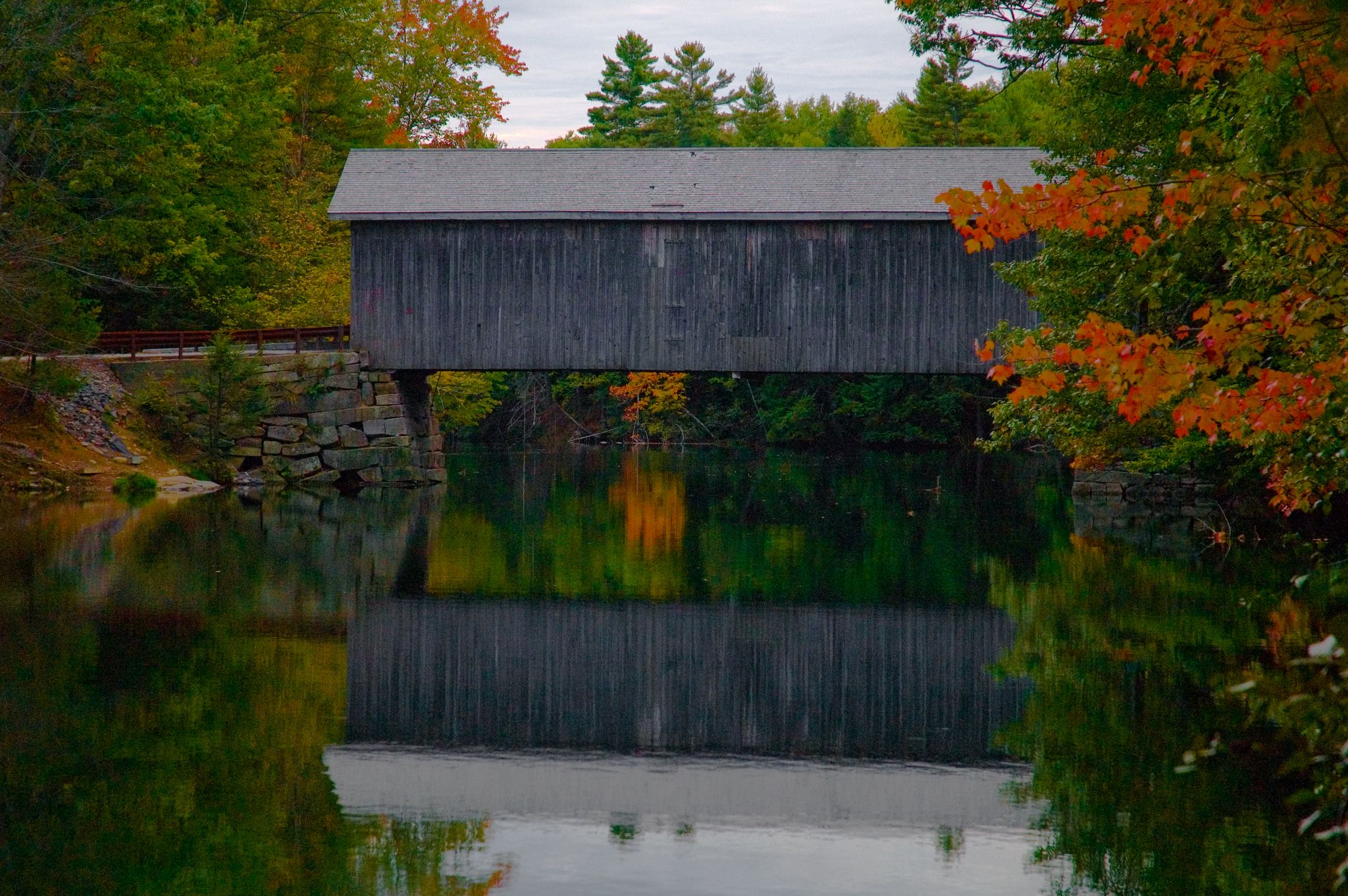 A covered wooden bridge spans calm water, surrounded by vibrant autumn foliage under an overcast sky. - River Road & Middle Jam Road loop via Babb's Bridge — Windham