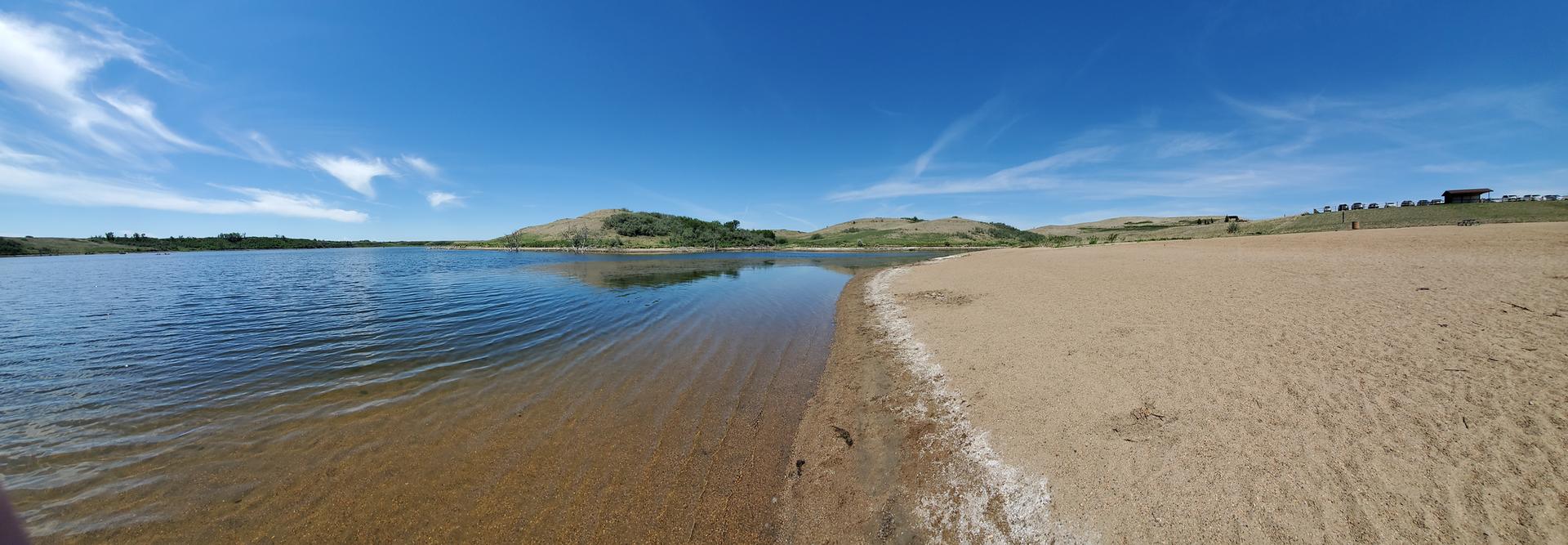 Panoramic view of a calm lake beside a sandy shore under a bright blue sky.