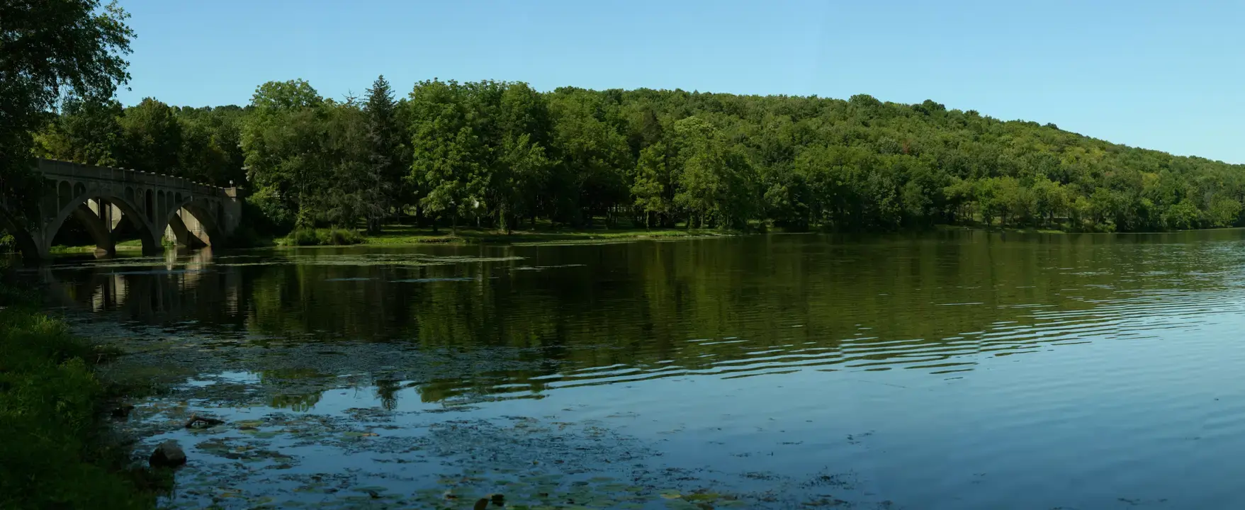 Stone bridge over a calm lake with green trees under a clear blue sky.
